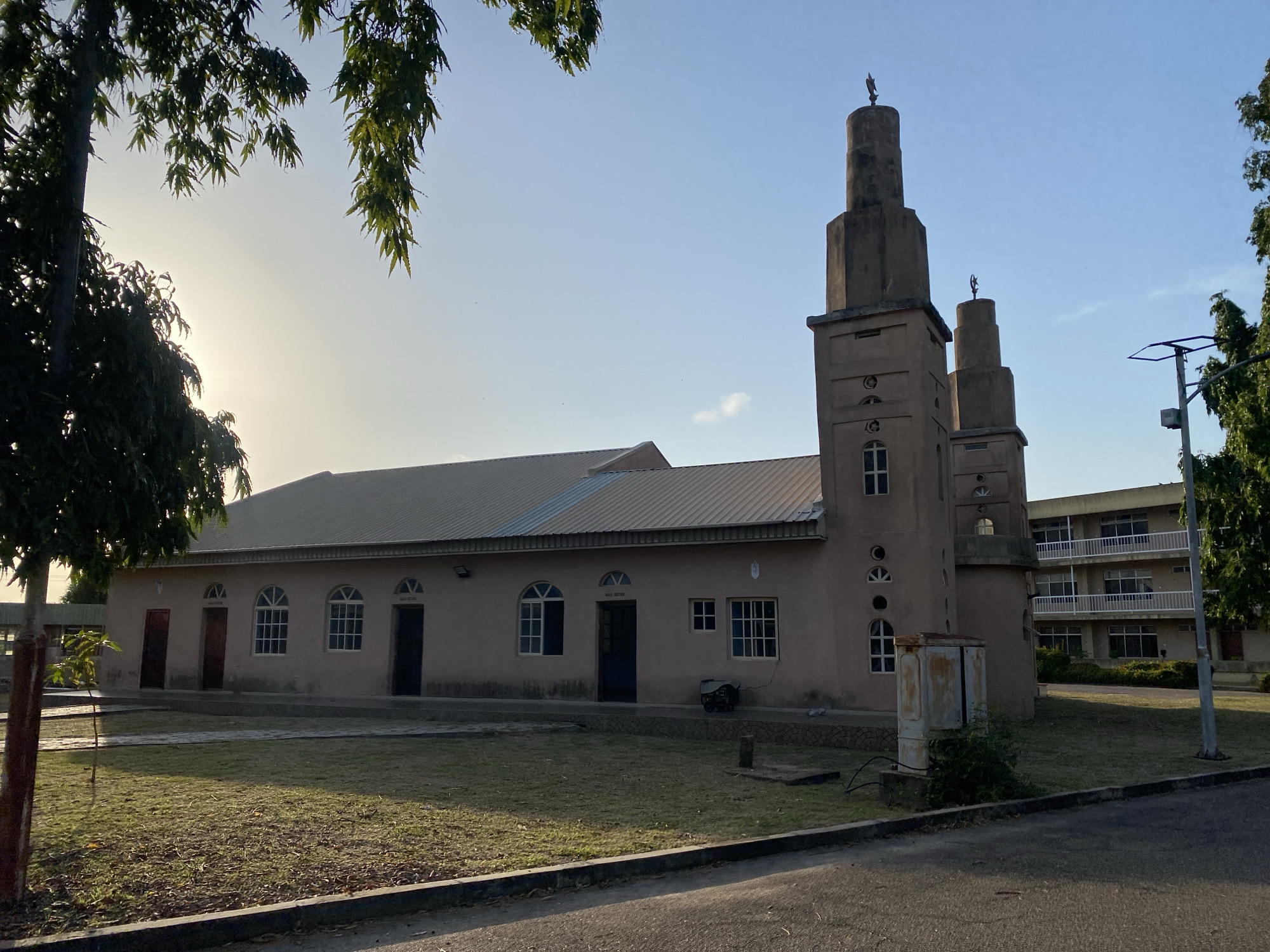 The image depicts a mosque in Tawpo, Nigeria. The mosque is a single-story building with a gabled metal roof. Attached to one side are two minarets, each topped with a crescent moon. The building's exterior is painted a light brown color. There are several windows and doors visible along the front facade of the mosque. The mosque is surrounded by a grassy area and a paved road. A multi-story building with balconies is visible in the background. A street light is present near the building in the background. Trees are scattered around the perimeter of the scene. The sky is pale blue, suggesting it may be early morning or late afternoon. No people are immediately visible in the image.