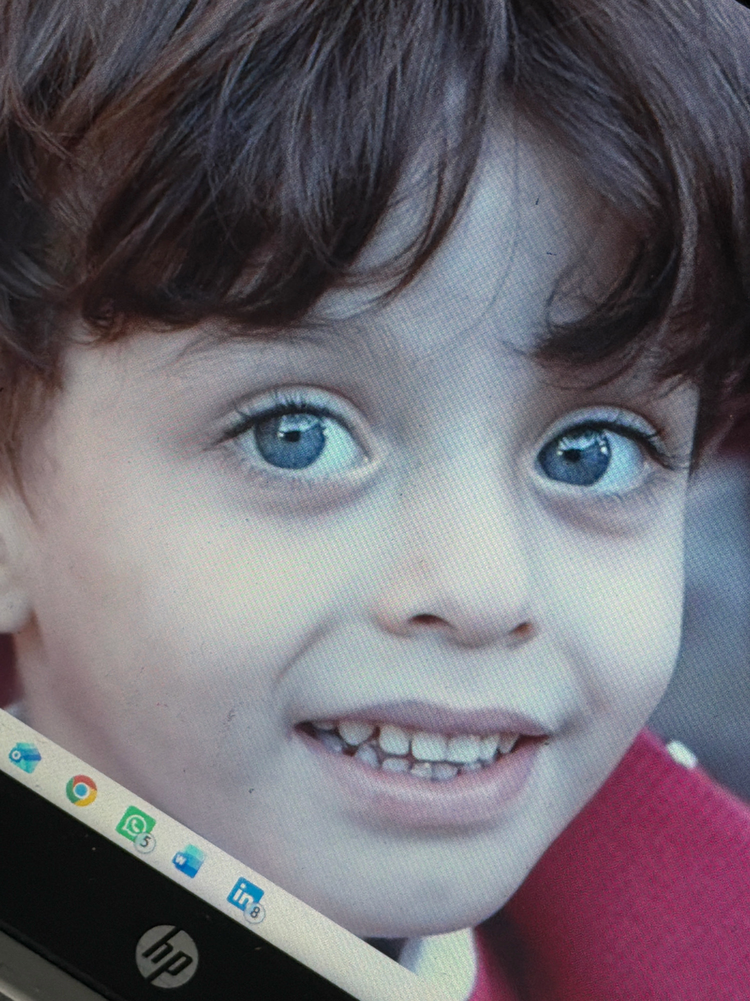 A close-up, eye-level photograph shows the face of a young child, likely under five years old, with fair skin, dark brown hair, and striking blue eyes. The child is smiling, revealing small, evenly spaced teeth. The child is wearing a pink garment on their right shoulder. The image appears to be a digital display, indicated by the presence of a laptop's taskbar at the bottom, showing icons for Google Chrome, WhatsApp, Windows, and LinkedIn, as well as an HP logo. The background is out of focus and nondescript. The photograph was taken indoors, likely in a home or office setting. There is no discernible information to determine the child's location in the city of Unknown, Unknown.