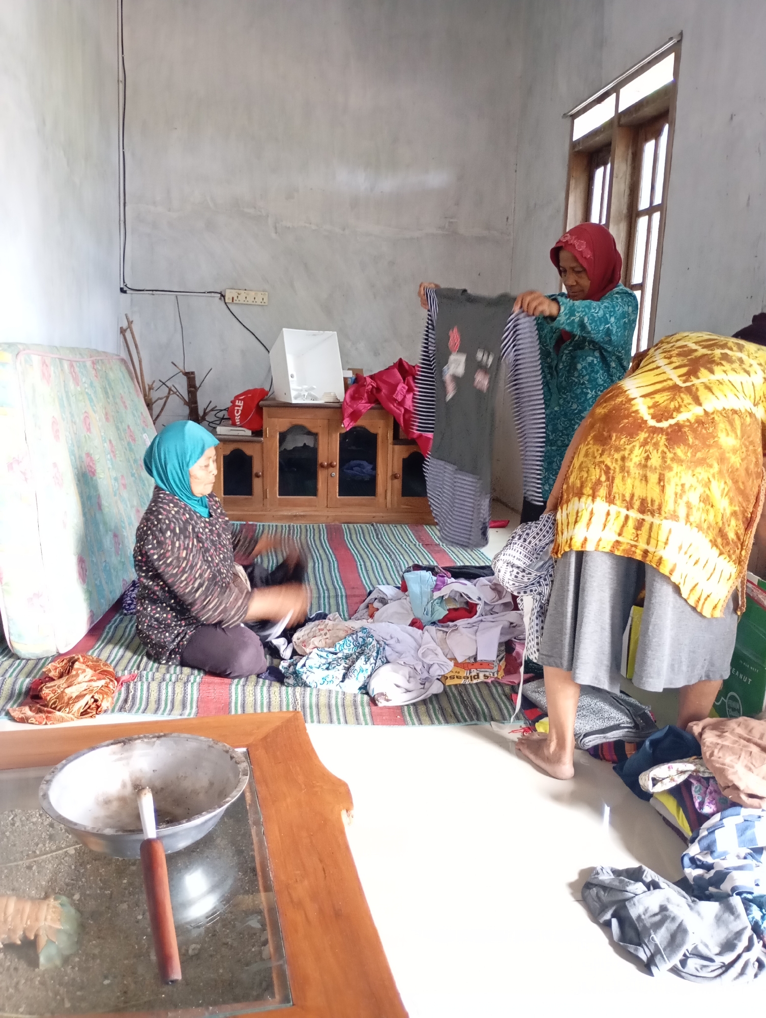 In Ketepung, Indonesia, a domestic scene unfolds in a modestly furnished room. Three women, dressed in traditional Islamic attire, are engaged in the task of folding laundry. Two women are kneeling on a striped mat on the floor, sorting and folding various garments. One woman, wearing a turquoise headscarf and a patterned top, is actively folding a grey garment with a design. To her right, another woman in a yellow and brown tie-dye patterned top and a grey skirt is standing, also interacting with the laundry. A third woman, wearing a light blue headscarf and a dark patterned top, is kneeling on the mat and appears to be in the process of folding clothes, with her hands blurred in motion. The room contains a mattress leaning against the wall, a wooden cabinet with glass doors, and a wooden coffee table with a glass top that holds an ashtray with a cigarette. Electrical outlets are visible on the wall, and natural light streams in from a window. The overall impression is one of everyday chores taking place in a simple, lived-in environment.