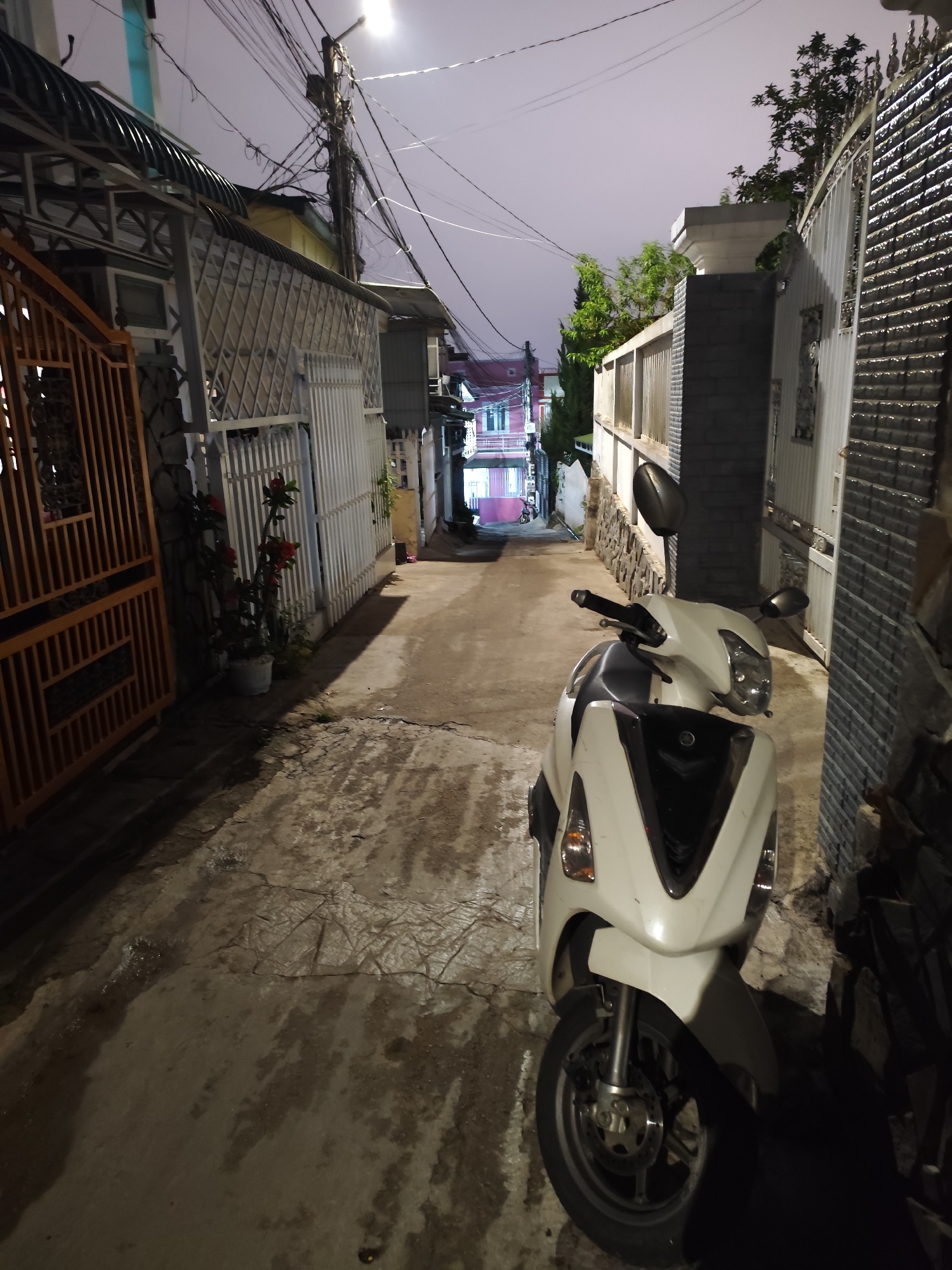 The image depicts a narrow alleyway in Da Lat, Vietnam, at night. The alley is paved with cracked concrete and slopes upwards into the distance. A white scooter is parked on the right side of the alley, partially blocking the path. Buildings line both sides of the alley, with a mix of gates and fences made of wood, metal, and brick. Overhead, a web of electrical wires crisscrosses the sky. A street light illuminates the alley, casting shadows along the walls and ground. In the background, a building with pink and blue accents is visible at the end of the alley. There are no people visible in the image. The scene is quiet and still.