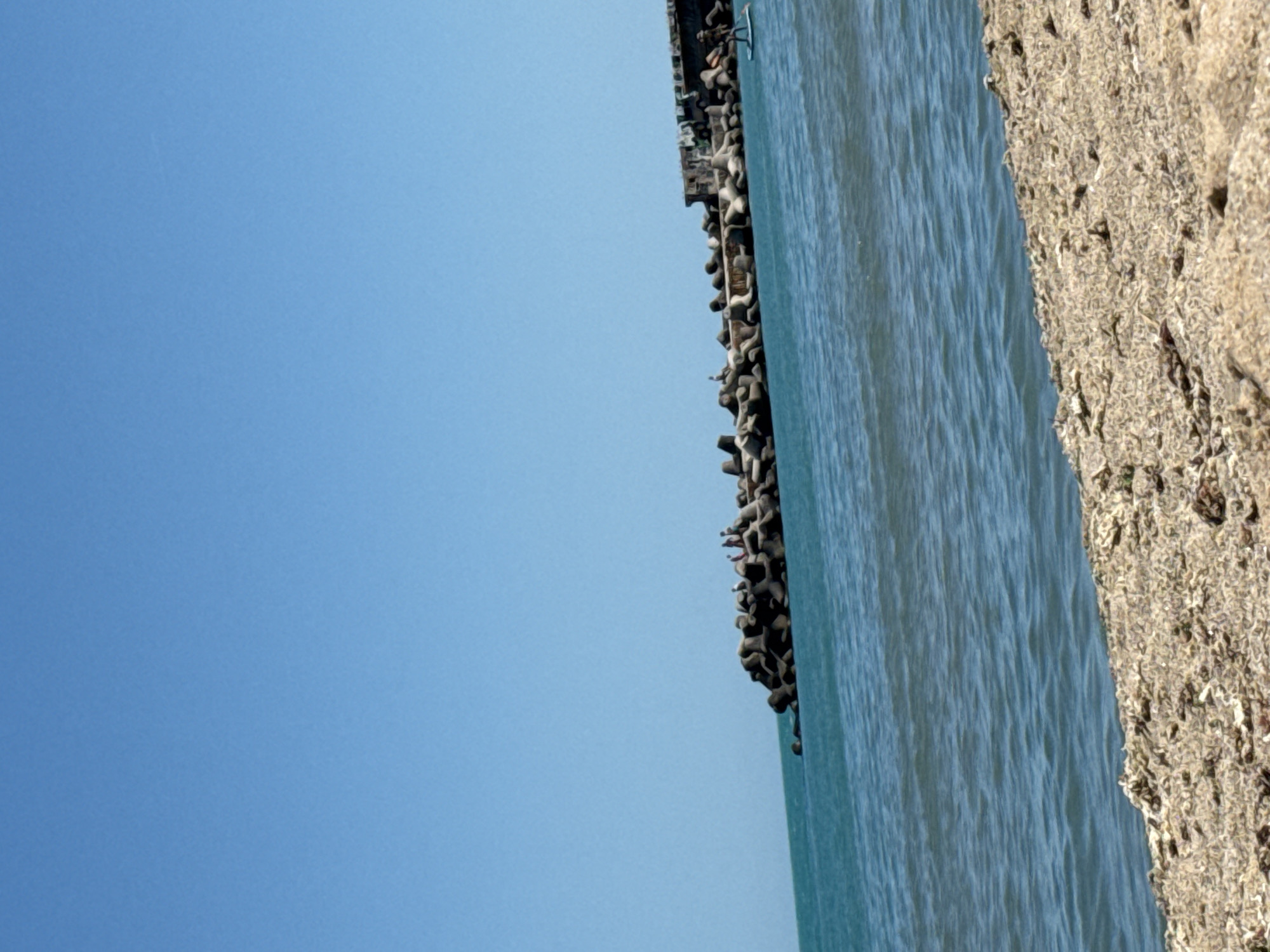 A bright, sunny day is depicted with a clear blue sky above the Black Sea. In the mid-ground, a breakwater made of large, dark grey concrete tetrapods extends into the turquoise water. Several small figures, appearing to be people, are visible standing on the breakwater, some of whom are interacting. To the right, a single person is standing on a paddleboard in the water near the breakwater. The foreground shows a sandy beach with scattered debris.