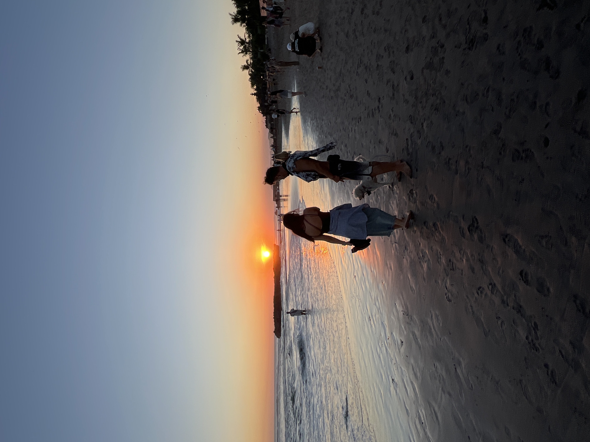 The image captures a scene at a beach, presumably in North Kuta, Indonesia, during sunset. The sky is a gradient of blue to orange, with the sun setting on the horizon. The beach is sandy with wet areas reflecting the sunset colors. Several people are present. A woman in a dark top and light blue skirt is standing near a man in a patterned shirt and shorts, who is holding a small white dog. Further down the beach, there are other people, some of whom appear to be sitting or bending down. Palm trees line the back of the beach. Footprints are visible in the sand. The scene is likely a popular sunset viewing spot.
