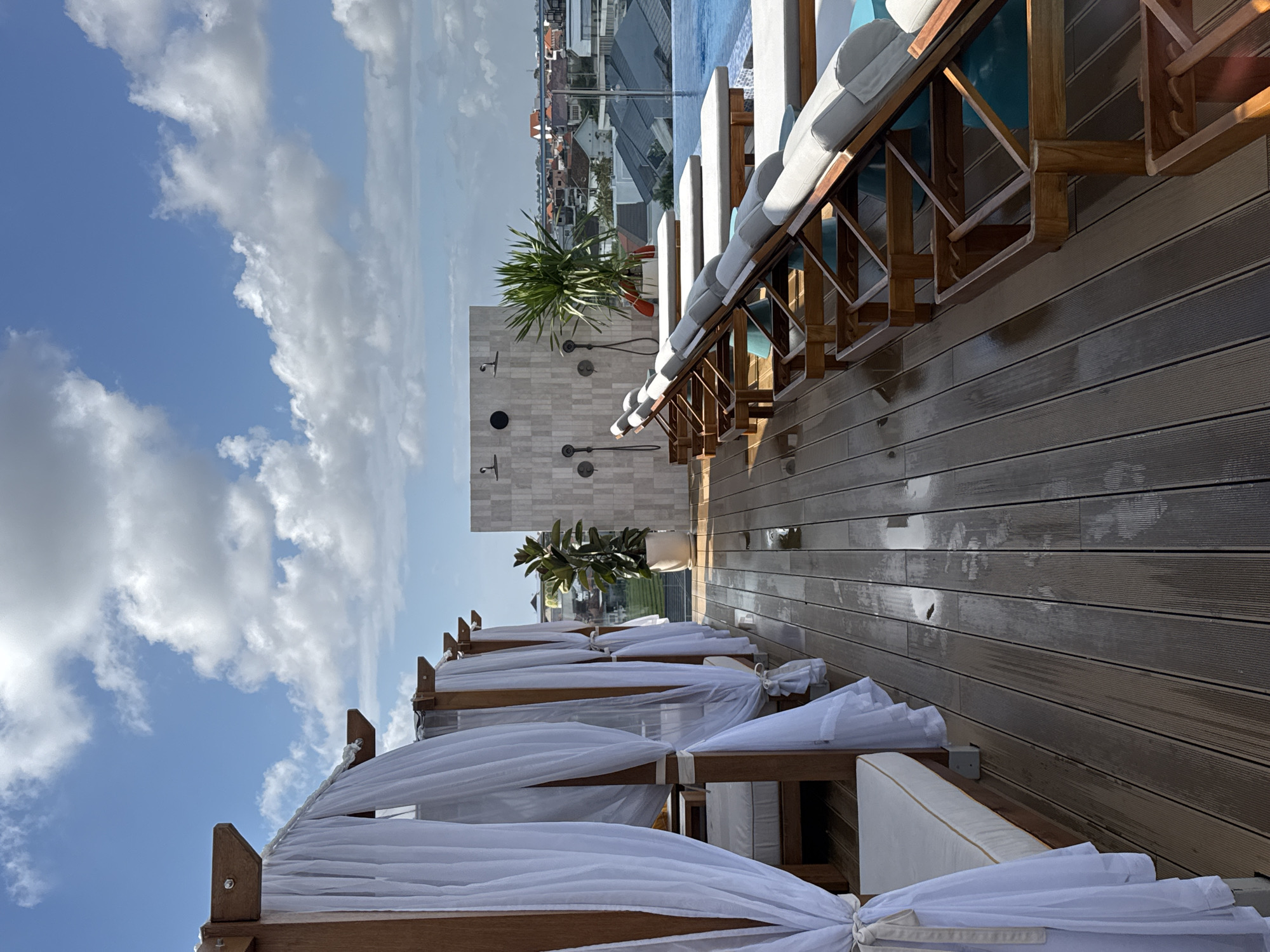 This image depicts a rooftop swimming pool area in North Kuta, Indonesia, under a partly cloudy sky. In the foreground, a wooden deck is wet with scattered water puddles. To the left, a row of white sheer curtains hang from wooden frames, obscuring cabana beds. To the right, several wooden lounge chairs with grey cushions are arranged, facing a clear blue swimming pool. In the middle ground, a tiled wall features multiple showerheads, and a large potted plant adds a touch of greenery. Beyond the pool, a cityscape with residential buildings and a distant view of the ocean can be seen. The scene appears to be unoccupied, suggesting a serene atmosphere.