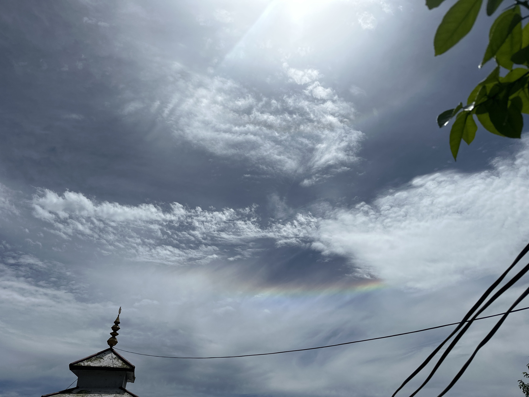 The image captures a sky scene in Bhagsu, India, dominated by a dramatic display of clouds and a rare atmospheric phenomenon. The sky is a canvas of gray and white clouds, with streaks of cirrus clouds creating a wispy texture. In the lower center of the frame, a faint rainbow-like arc, possibly a circumzenithal arc or a tangent arc, is visible, its colors muted against the cloudy backdrop.
In the foreground, the peak of a building, likely part of a temple or religious structure, is visible on the lower left. It features a tiered, golden spire atop a white structure with a red-tiled roof. Dark electrical wires stretch across the middle ground, casting shadows against the sky. On the upper right, a cluster of green leaves from a tree dangles into the frame, providing a natural element against the man-made and atmospheric features. The overall impression is a serene yet awe-inspiring moment of natural beauty observed from a specific location in Bhagsu, India.