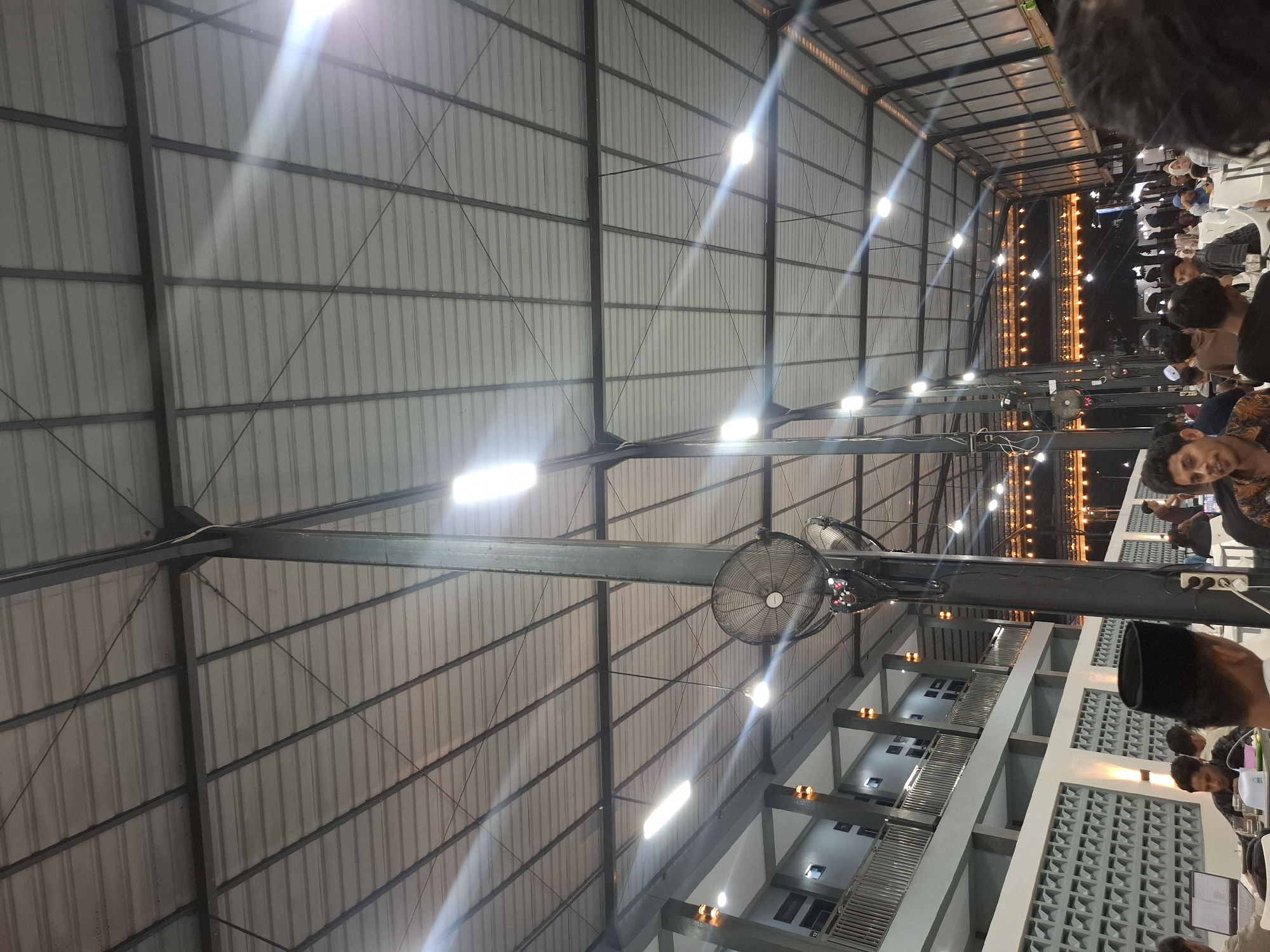 The image depicts an indoor gathering at night, likely an event or a restaurant, located in Kampongbaro, Indonesia. The ceiling is a corrugated metal roof supported by a steel frame structure. Numerous fluorescent lights illuminate the space, casting long beams and reflections across the metal surface. Large industrial fans are mounted on vertical support poles. In the foreground, several men are seated at tables, some engaged with laptops or food. One man in the center foreground wears a black peci hat. In the background, more people are visible, and further back, decorative string lights create a warm glow. The architecture suggests a covered outdoor or semi-outdoor space with a second-story balcony or walkway visible on the left. The overall atmosphere is one of a communal event or dining experience.