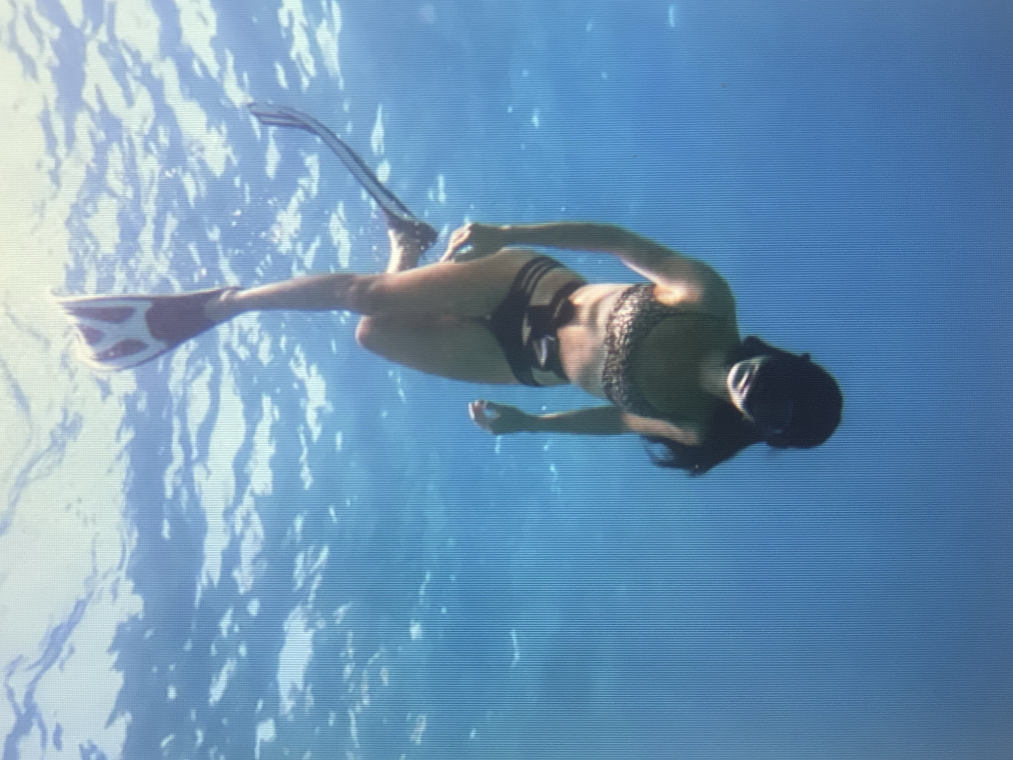 An upside-down, full-body shot shows a person wearing a black snorkel mask and fins, with a leopard-print bikini top and bottoms, and a black band around their waist. The person is submerged in clear blue water, with sunlight filtering through the surface from above. The background is a solid blue expanse of water with ripples on the surface.