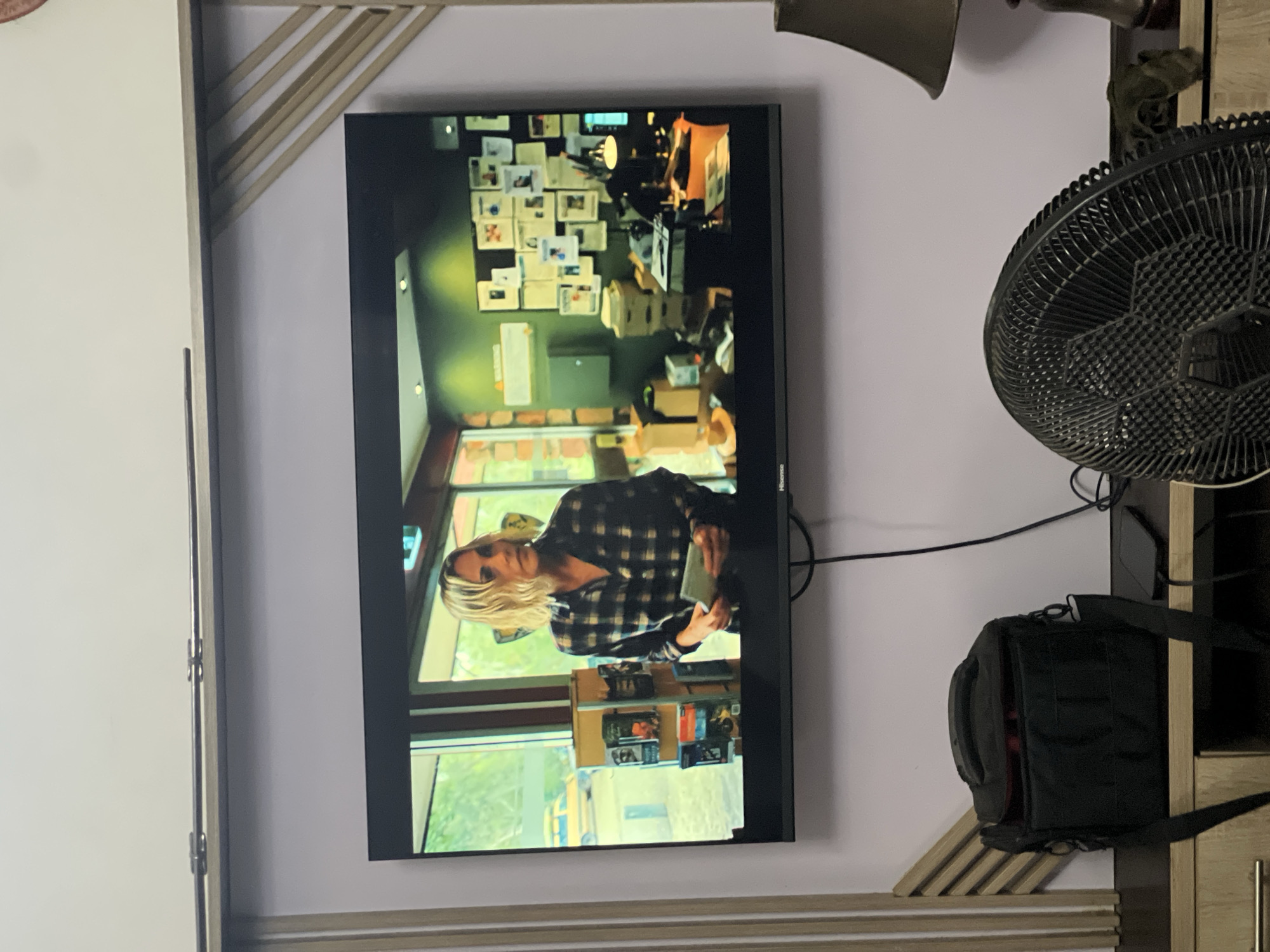 A television is mounted on a wall, displaying an image of a blonde woman in a plaid shirt. The scene on the screen appears to be set in a building with numerous documents and items on shelves visible in the background. To the right of the television, there's a black fan and a black bag. The wall is white. The scene is indoors. The location in the city of Abuja, Nigeria cannot be determined from the image.