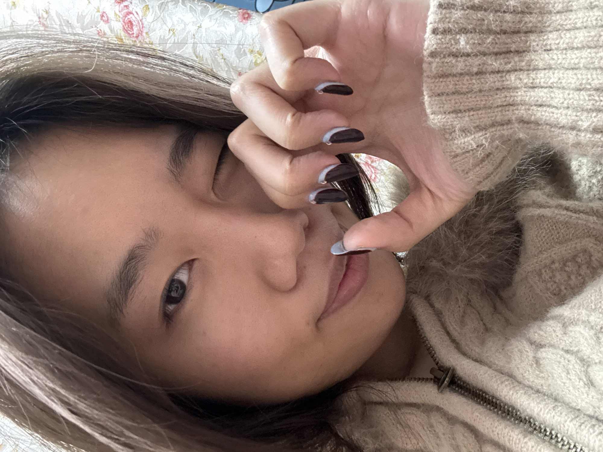 A close-up image displays a female individual, appearing to be of East Asian descent, located in Shapingba District, China. The subject's left eye is open, while her right eye is closed in a wink, accompanied by a slight smile. Her light brown hair partially covers the right side of her face. Her right hand is raised, with the index finger touching her lower lip or chin. The fingernails on her right hand are painted with a dark base color, possibly black or dark brown, and have light blue or white tips in a French manicure style. A silver ring is visible on her right ring finger. She is wearing a light beige or cream-colored knitted garment, likely a sweater or jacket, featuring a cable-knit pattern on the body and ribbed cuffs. A silver zipper with a pull tab is visible on the front, and a light-colored faux fur trim is present around the collar or neckline. The background is out of focus, revealing a light-colored textile pattern with prominent pink roses and green leaves.