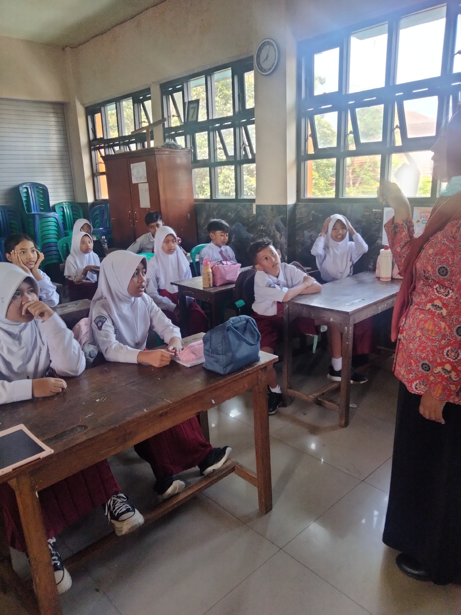 In Trawas, Indonesia, a classroom scene unfolds with a teacher, partially visible on the right side of the frame, engaging with a group of students. The students, dressed in white shirts and maroon skirts or pants, are seated at wooden desks, some looking towards the teacher and others at their materials. A few students are wearing headscarves. The classroom appears to be well-lit with natural light streaming through large windows that offer a view of trees and the sky. Desks, chairs, a cabinet, and stacks of plastic chairs are visible elements of the environment. The overall impression is one of a typical school day in session.