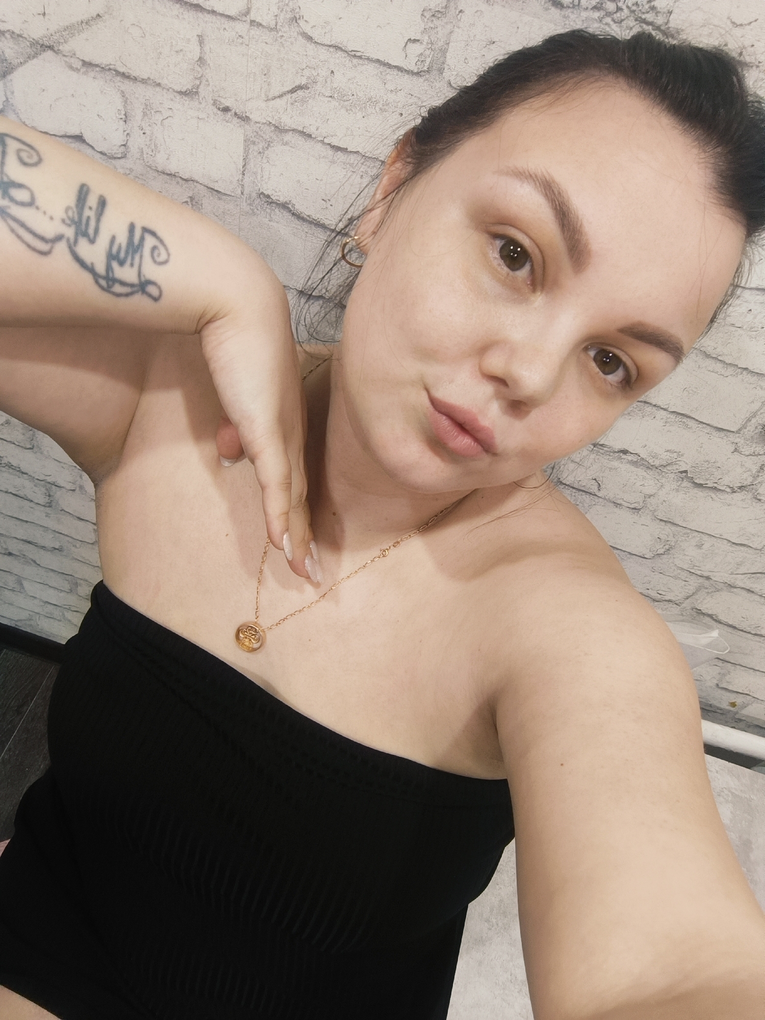 A close-up studio shot depicts a fair-skinned female subject wearing a black, strapless top and a gold necklace with a round pendant. The woman’s left arm is raised, displaying a tattoo on the upper arm. Her facial features include dark eyebrows and brown eyes. The background consists of a light-colored brick wall. Location is unknown.