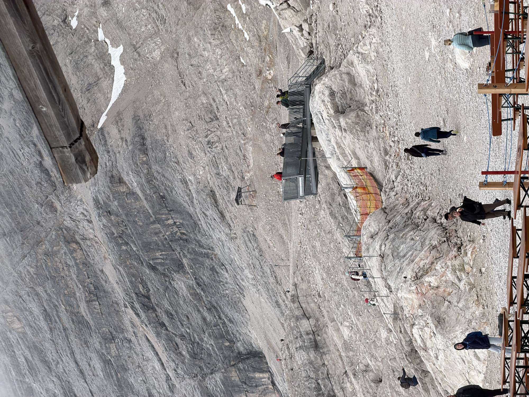This image captures a scene at the Zugspitze, Germany's highest peak, near Garmisch-Partenkirchen. The foreground features several wooden picnic tables, some with benches, set up on a gravelly surface. A portion of a wooden overhang is visible in the upper right. In the middle ground, a viewing platform with metal railings and stairs is occupied by a group of people. Further up the rocky, steep mountainside, other individuals are seen walking and navigating the terrain, with some holding onto safety ropes. The mountainous environment is characterized by sparse vegetation, large rock formations, and patches of snow. The sky is overcast.