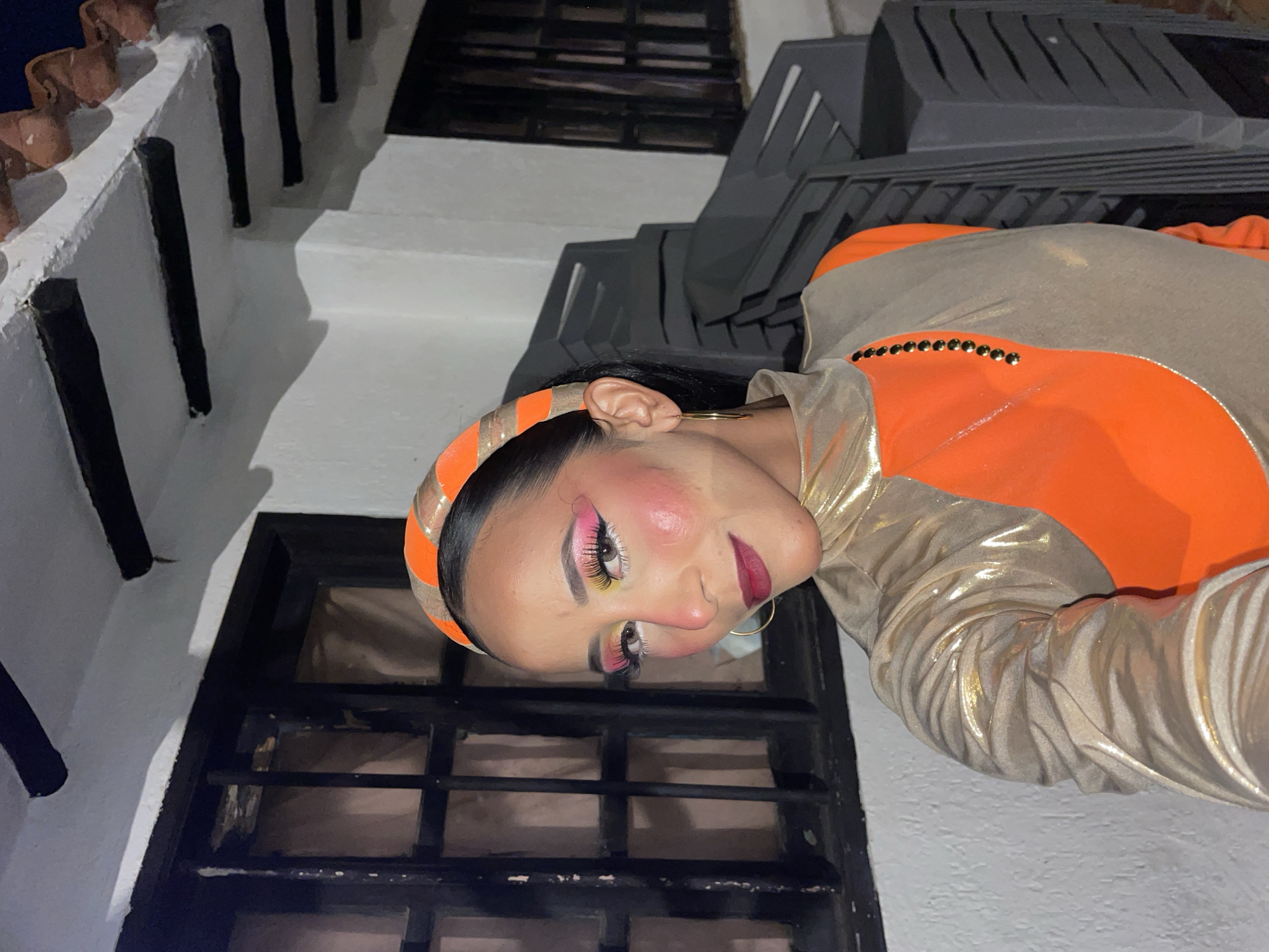 A woman poses for a selfie, her face angled towards the camera, in an outdoor setting at night. She wears a striking outfit consisting of a gold, high-necked top with a contrasting orange overlay. Her makeup is dramatic, featuring vibrant pink and yellow eyeshadow, rosy blush, and dark lipstick. She accessorizes with a gold headband with orange accents and a simple gold hoop earring. Behind her, dark-colored chairs are stacked, and a building with dark-trimmed windows and a white, textured facade is visible. The roofline features dark beams and terracotta tiles. The scene suggests an event or gathering, possibly in a courtyard or patio area. The woman's attire and makeup indicate a festive or theatrical occasion. The location is specified as Porlamar, Venezuela.