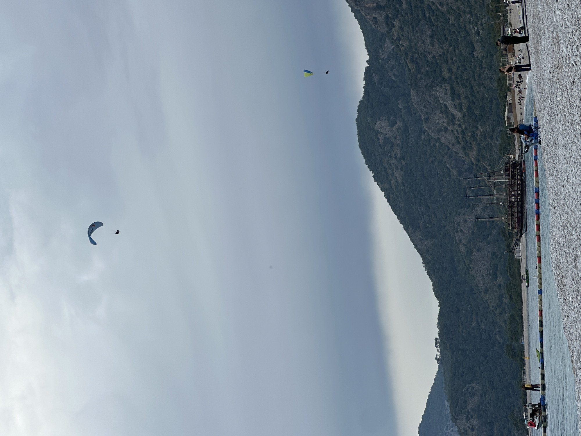 The image captures a serene coastal scene in Fethiye, Turkey, under an overcast or hazy sky. The perspective is from a pebbly beach looking out towards the sea and a large, forested mountain range.
In the mid-ground and background, a towering, dark green, tree-covered mountain dominates the right side of the frame. Below the mountain, calm, light blue water stretches out, separated from the foreground beach by a line of colorful buoys, likely marking a swimming area.
Two paragliders are visible in the expansive sky on the left. One, closer to the viewer, is blue and white, with a person clearly visible suspended beneath it. Further in the distance, another paraglider, yellow and blue, also carries a person.
On the pebbly beach in the foreground and along the shoreline, several people are present. One individual is seated in a blue beach chair near the water's edge, while others stand or stroll further along the shore. A prominent feature near the water is a large, dark-hulled, multi-masted vessel resembling a traditional pirate ship, anchored or docked by the shore.
The time of day appears to be daytime, with diffused light indicating either an overcast sky or a hazy atmosphere. No discernable text is visible in the image.