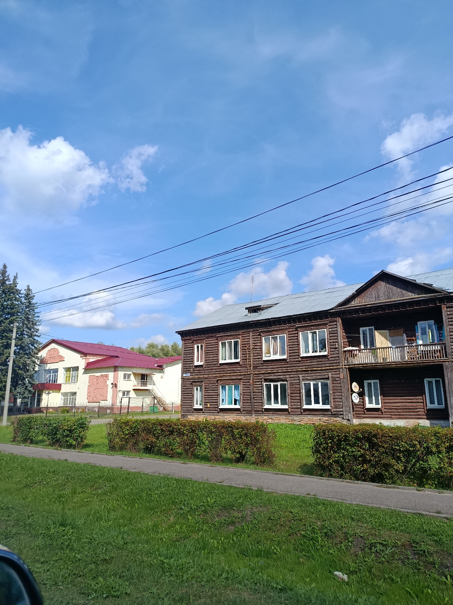 The image depicts a street scene in the city of Vereya, Russia. In the foreground, a section of a grassy area and a paved path are visible, suggesting a public space or sidewalk. Beyond the path, a neatly trimmed hedge runs horizontally across the scene, separating the path from two buildings.
The building on the right is a two-story wooden structure with a dark brown, log construction. It features multiple white-framed windows on both floors. The upper floor has a covered balcony with railings, on which some items are placed, and a white-framed door leading onto it. Two circular satellite dishes are mounted on the exterior wall.
To the left of the wooden building is a lighter colored building with a prominent red roof. This building appears to be of a more modern construction, with a light yellow facade and decorative pink relief panels on its exterior walls. It has several white-framed windows and a visible external staircase leading to a small landing on the side. Tall evergreen trees stand to the far left of this building.
The sky above is a bright blue with scattered white clouds and several dark power lines crisscrossing the upper portion of the image. The overall impression is a view of residential or communal buildings in a Russian town on a clear day.