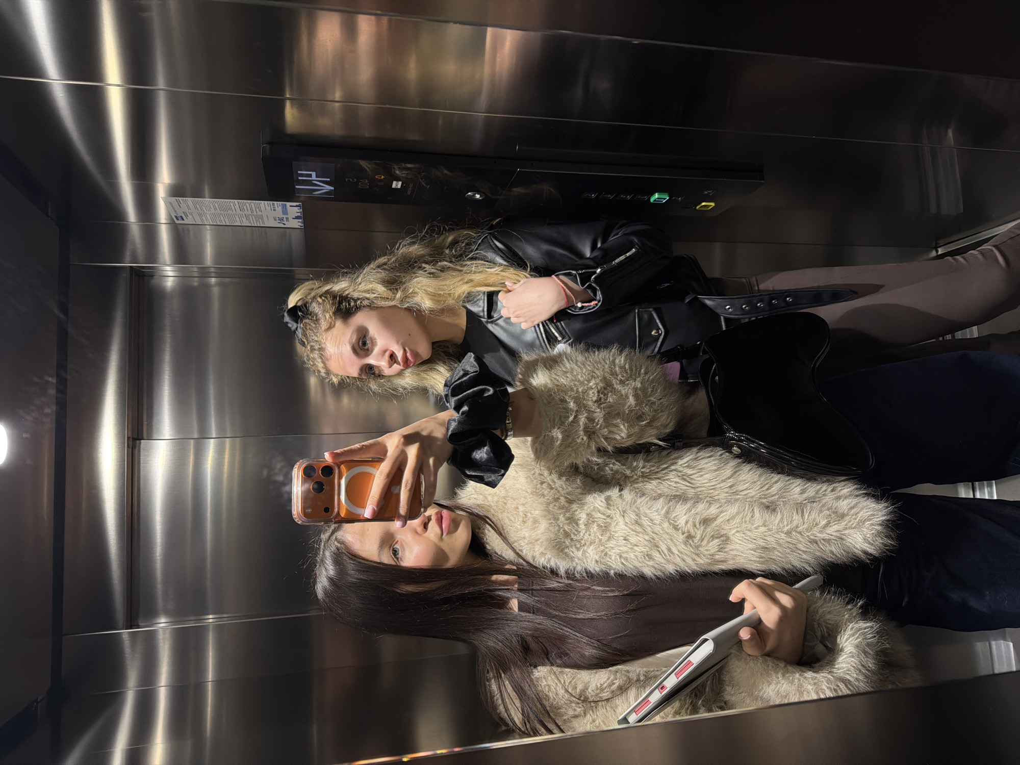 Two young adult females are captured in a mirror selfie within a modern, polished stainless steel elevator in Sofia, Bulgaria. The female on the left, displaying long dark hair, wears a dark top, a light beige faux fur coat, and dark trousers, holding a grey tablet or laptop. The female on the right, with wavy blonde hair secured in a black scrunchie, wears a black leather jacket, a dark top, and brown leggings, carrying a black patent leather handbag. She holds an orange-cased smartphone with her right hand to take the selfie; both individuals are looking towards the device, with the selfie-taker exhibiting a puckered lip expression. Her left hand rests near her chest. An elevator control panel on the right wall features a black display indicating "NV", floor "2", and "004", alongside green and yellow directional buttons. A white informational sticker displaying "JAS" (referencing the Bulgarian elevator company JAS-VRLD) and Cyrillic text is visible on the wall above the control panel, confirming the location as Sofia, Bulgaria. The elevator interior is uniformly lit by an overhead light source.