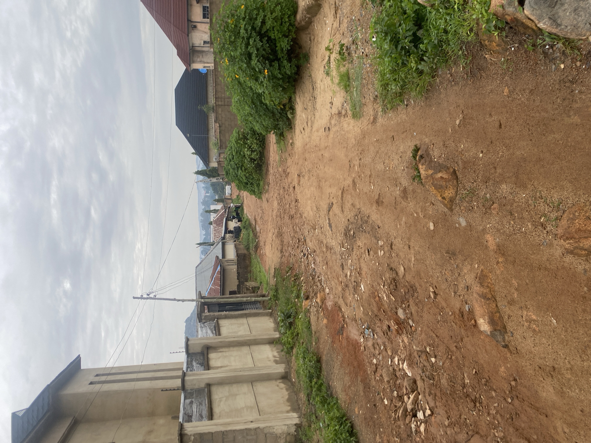 The image depicts a dusty, unpaved dirt road in a residential area of Jos, Nigeria. The road surface is a mix of red-brown soil, scattered stones, and some debris. On the left side of the road, a tall, light-colored concrete wall with a dark metal gate runs along the property line. Beyond the wall, a portion of a building with a dark-tiled roof is visible. In the distance, several houses with various roof styles and colors, including dark grey and reddish-brown tiles, are scattered among green vegetation. Tall trees and lush green bushes, some with yellow flowers, line the sides of the road and the properties. Overhead, power lines stretch across the scene, supported by utility poles. The sky is overcast with clouds, suggesting a diffused light or possibly an impending rainfall. The overall scene conveys a sense of a developing or less urbanized neighborhood within Jos.
