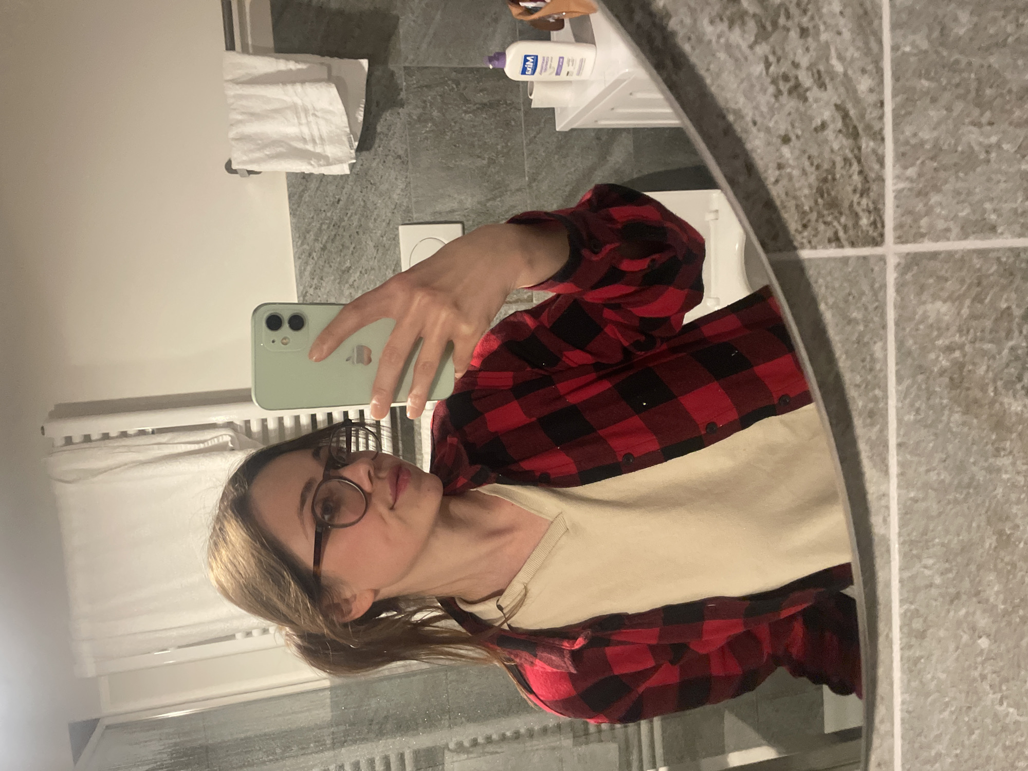 A woman with light brown hair pulled back in a low ponytail, wearing round glasses and a red and black flannel shirt over a cream-colored t-shirt, is taking a selfie in a bathroom mirror. She is holding a light green iPhone with both hands. The bathroom has grey tiled walls. A white towel is hanging on a rack, and a bottle of lotion or soap sits on a shelf above the sink. A radiator is visible in the lower part of the frame. The location is possibly Sappada / Plodn / Sapade, Italy.