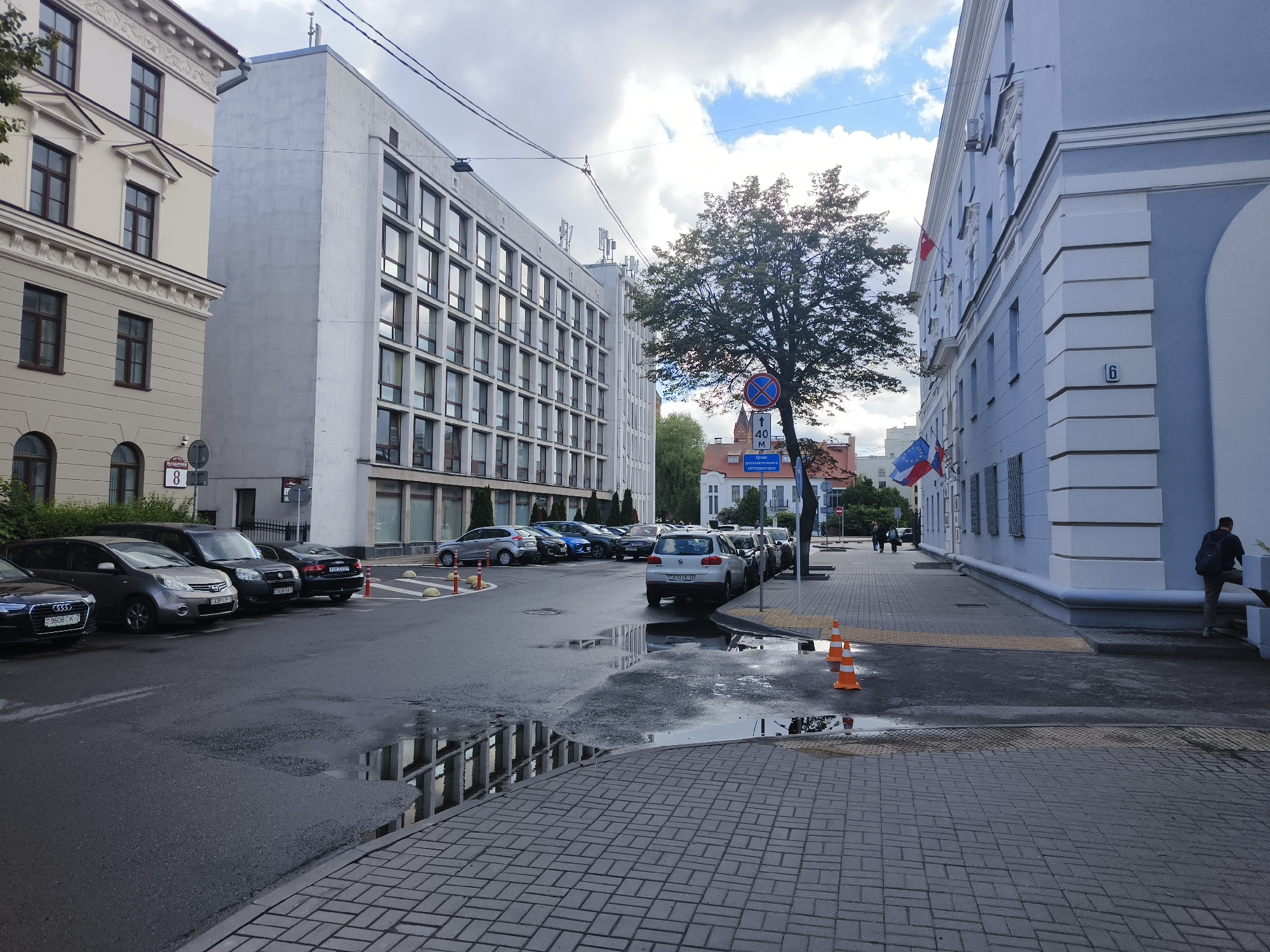 The image depicts a street scene in Minsk, Belarus, with a light overcast sky suggesting a cloudy day. Several cars are parked along the wet asphalt road, reflecting the surrounding buildings and sky in the puddles.
On the left, a light-colored, multi-story building with numerous windows stands prominently. To its left, another building, lighter in color and with more ornate architectural details on its upper floors, is partially visible. A street sign with the number "8" is affixed to this building.
The center of the image features a larger, modern building with a repetitive grid of windows. A large tree with green foliage stands in front of it, casting shadows. A circular traffic sign with a red border and blue center, indicating no entry for vehicles, is visible near the tree, along with a sign indicating "40 M" and some Cyrillic text.
To the right, a light blue building with white architectural accents dominates the foreground. A number "6" is visible on its facade. Two flags, one red and white, and another with blue, white, and red stripes, are displayed on this building. A person with a backpack is walking away from the camera on the sidewalk in front of this building.
The overall scene suggests a typical urban environment in Belarus, with a mix of older and newer architecture, parked vehicles, and signs of recent rain. The presence of the flags hints at a possible official or governmental building. The time of day appears to be late afternoon or early evening, given the lighting conditions.