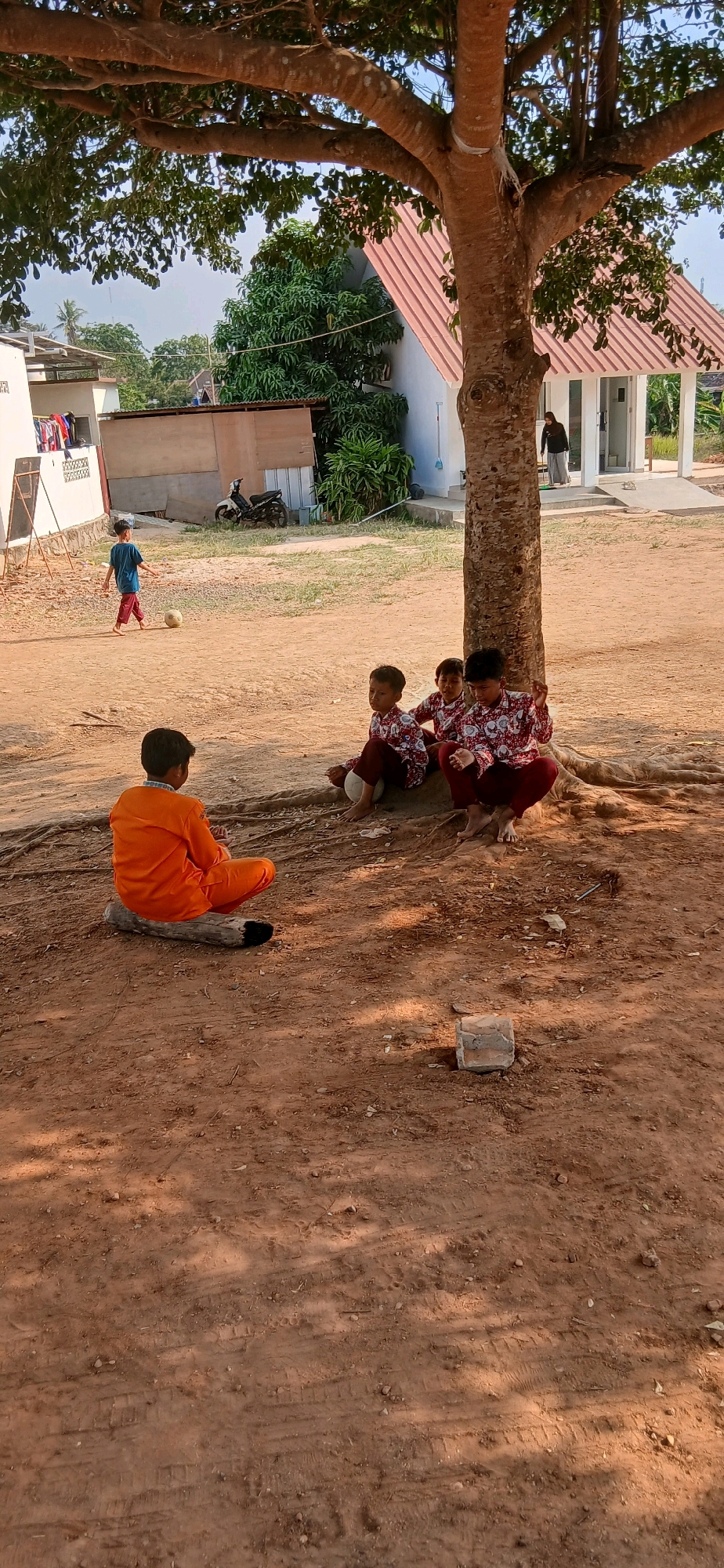 In the city of Metro, Indonesia, a dirt area is depicted under a large tree. Five young boys are present. One boy, wearing an orange shirt and pants, sits on a log facing away from the camera. Three other boys, dressed in matching red and white patterned shirts and maroon pants, sit near the base of the tree. One of these boys has a white ball at his feet. A fifth boy, wearing a blue shirt and maroon pants, is a short distance away, kicking a soccer ball. In the background, there are several buildings, including a white structure with a red roof and another building with a corrugated roof. A motorcycle is parked near one of the buildings, and laundry hangs on a clothesline. The ground is sandy and uneven, with some debris, including a brick, scattered around. The scene appears to be an outdoor recreational area or schoolyard.
