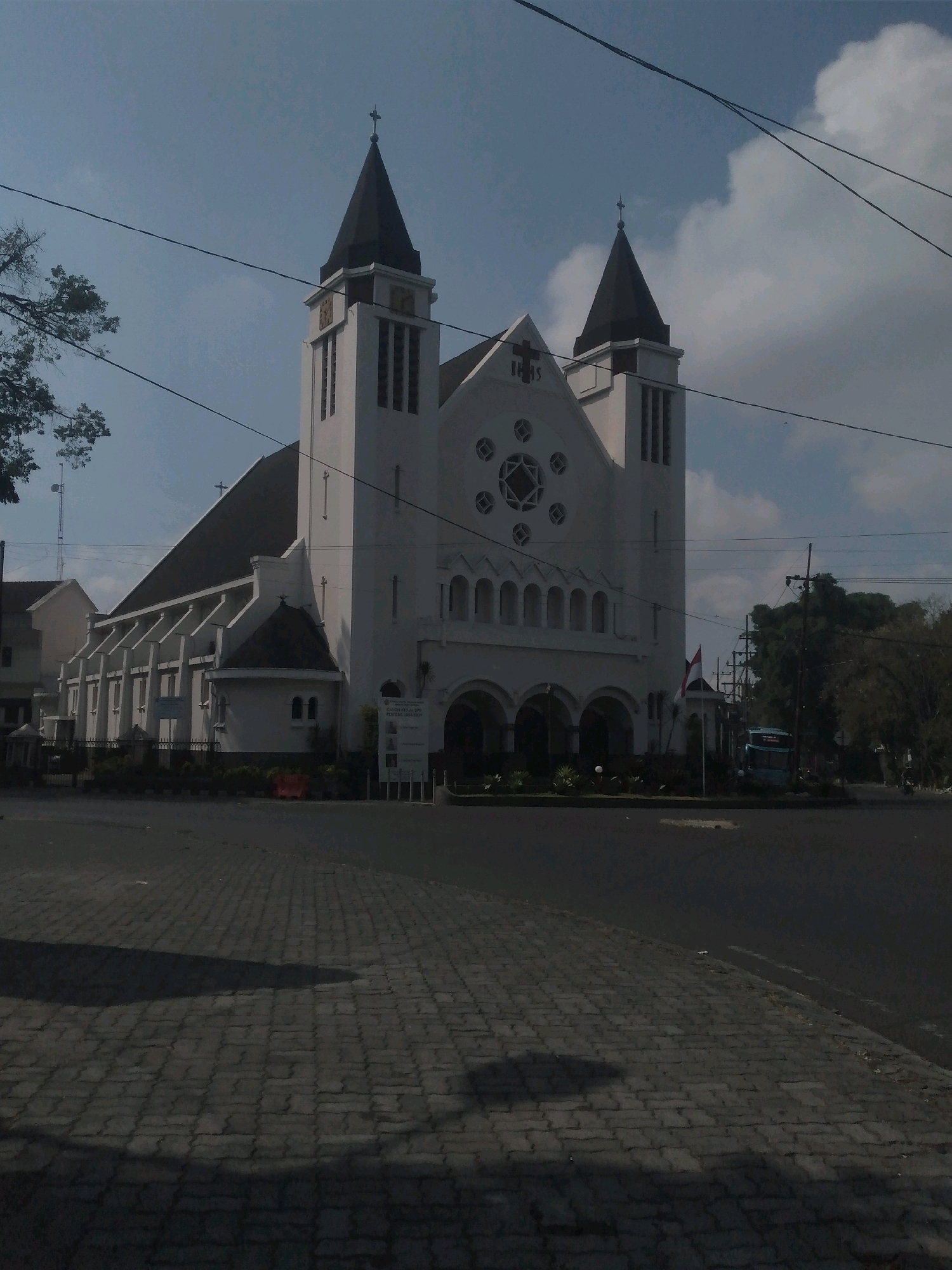 The image shows the Immanuel Church in Malang, Indonesia. The church is a large, white building with two spires, a peaked roof, and arched windows. It is located on a street with a paved sidewalk and a road. There are trees and power lines in the background. A blue bus is parked on the side of the road. The sky is partly cloudy. The image is taken from a low angle, looking up at the church.