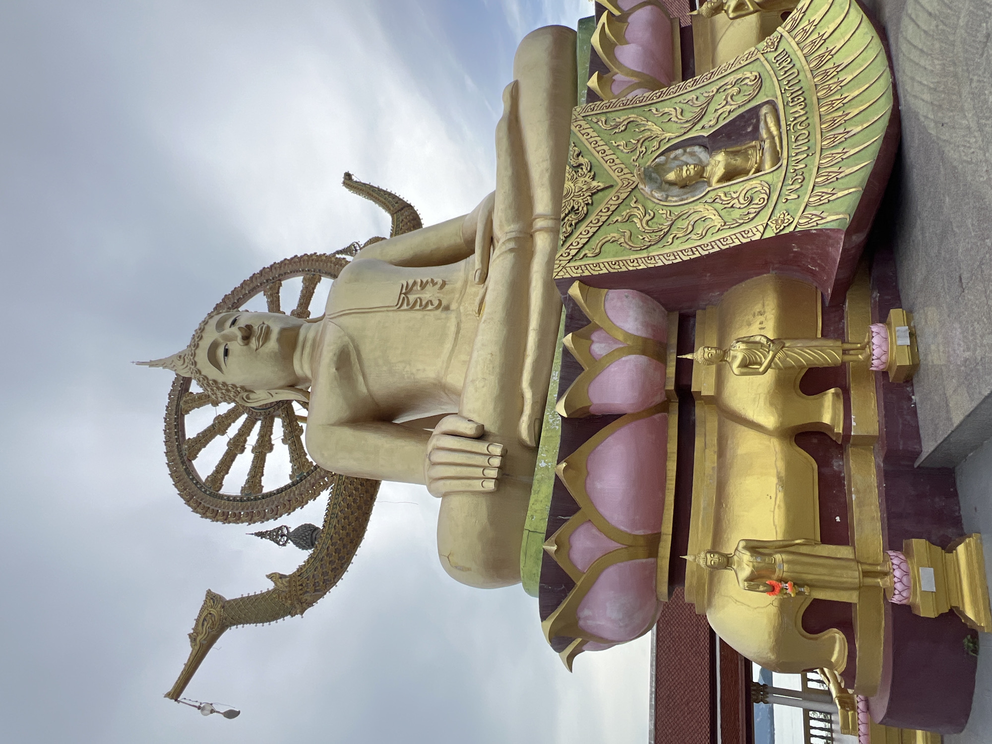 A large, golden Buddha statue sits in a meditative pose atop a multi-tiered, lotus-shaped pedestal. The statue is adorned with intricate details, and a Dharma wheel is positioned behind its head. To the left of the Dharma wheel, a golden dragon-like sculpture extends outwards, with a small bell or charm hanging from its snout. The base of the statue is decorated with smaller golden figures, one of which holds a floral garland. A large, ornately decorated green and gold banner with an inscription and a smaller Buddha image is draped across the front of the pedestal. The scene appears to be outdoors, with a cloudy sky in the background. This is a view of the Big Buddha at Baan Plai Laem Temple in Koh Samui, Thailand, a prominent landmark and popular tourist destination.