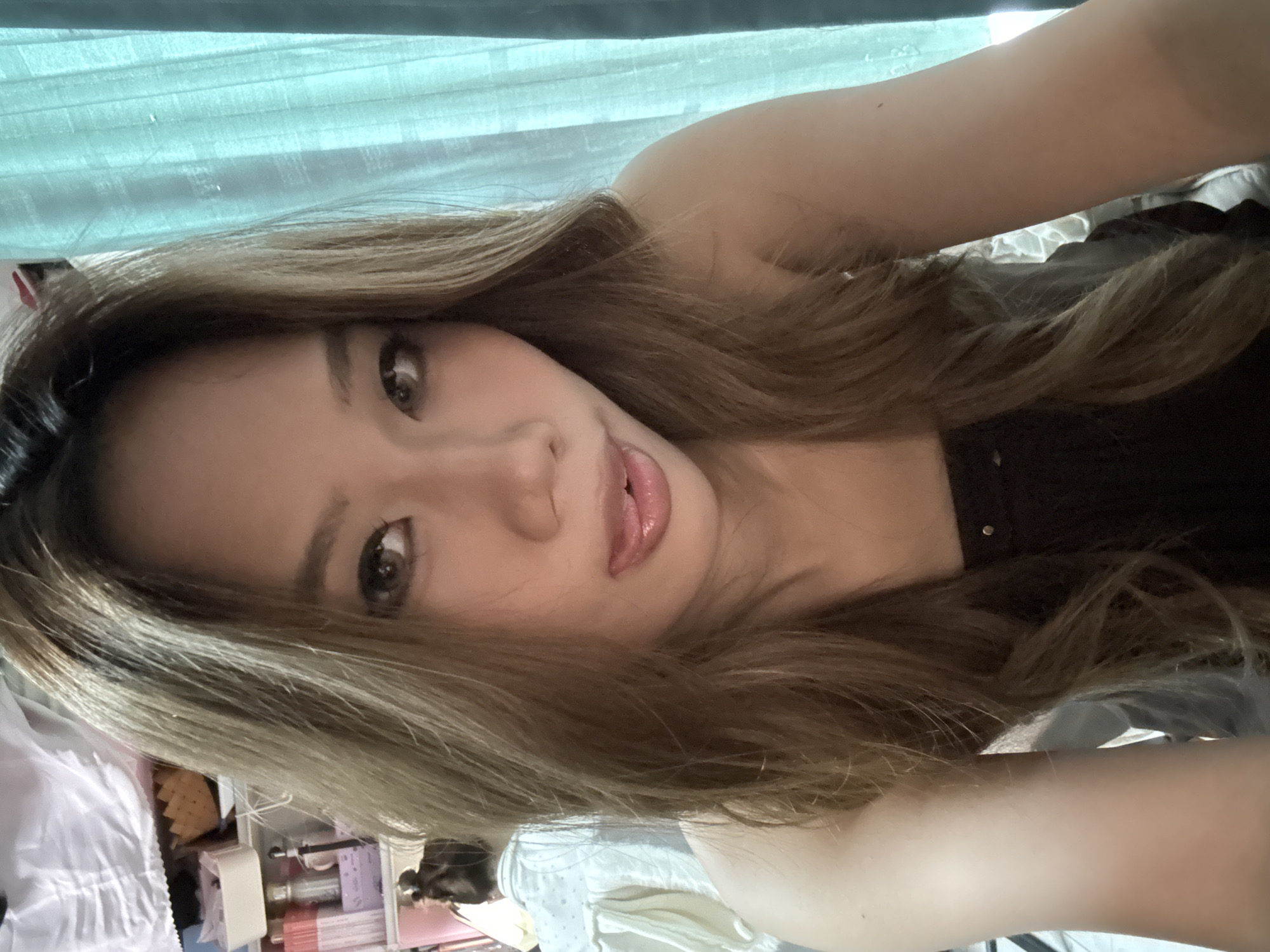 A young woman with long brown hair and brown eyes is taking a selfie. She is wearing a dark-colored top. The background includes a light blue curtain, a white sheet, and some items on a shelf. The location appears to be indoors. The city information is not detectable.