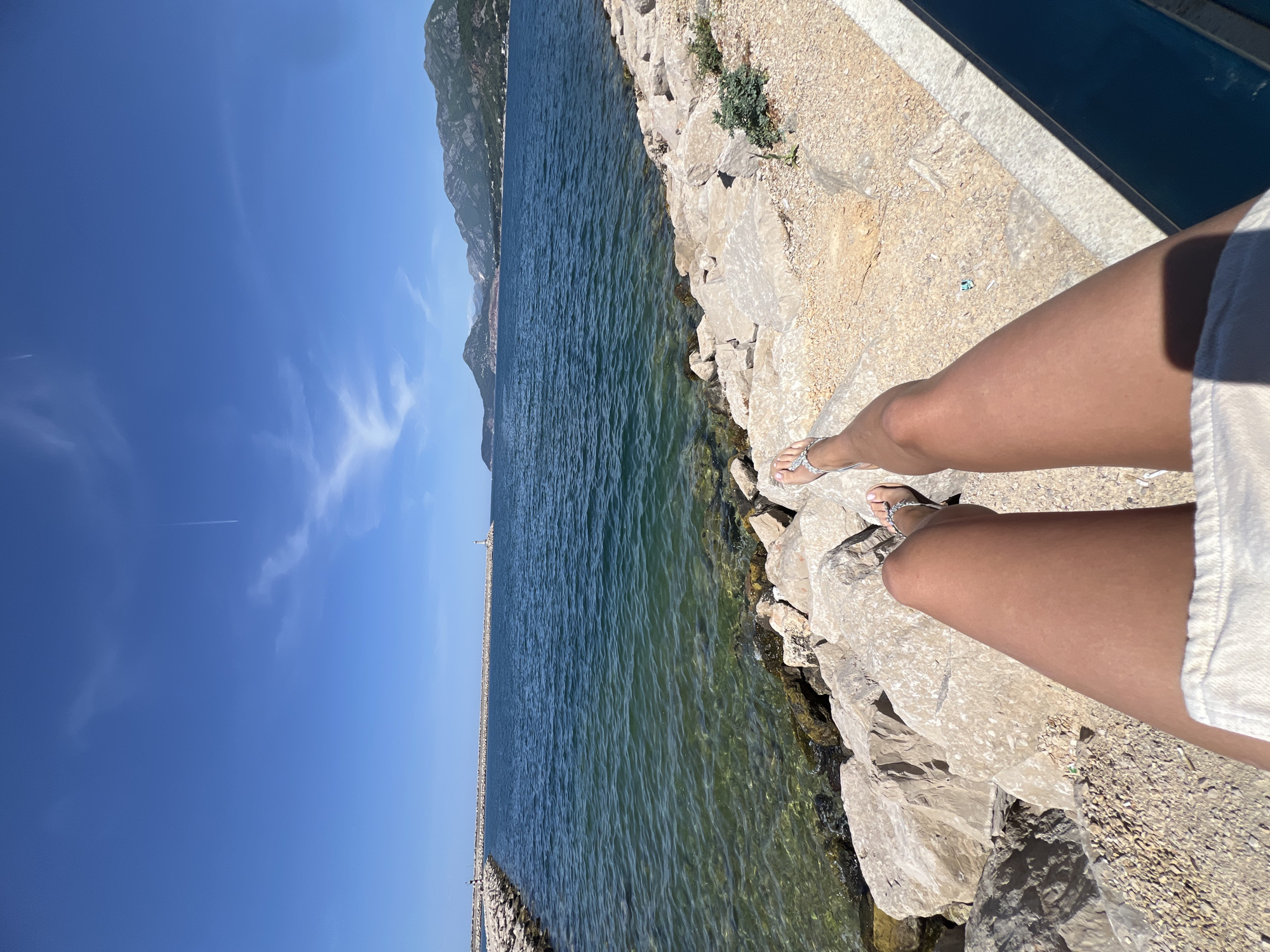 A long-angle eye-level outdoor shot captures a section of the shoreline in Šušanj, Montenegro. The perspective is oriented toward the left side of the frame. A person is observed standing on the rocky shoreline, their legs visible on the right side of the image. They are wearing silver sandals and a white garment. The shore is composed of light-colored rocks and small patches of vegetation.
The water extends from the near foreground toward the distant mountain range. The water appears clear with a blue-green tint and ripples on the surface. A long pier or breakwater, lined with posts and structures, divides the frame in the distance. The sky is a clear blue with sparse clouds. The overall lighting suggests a sunny day.