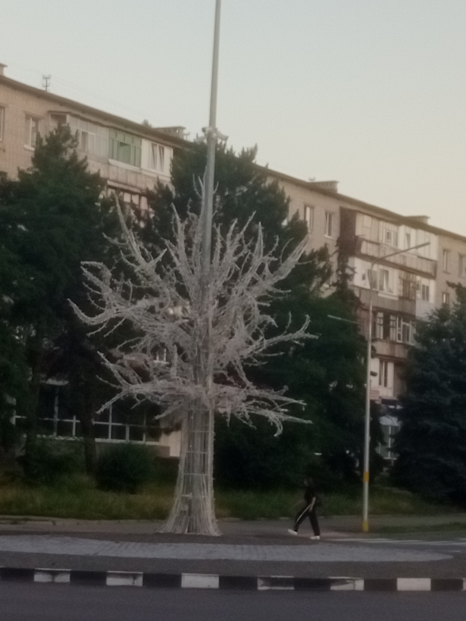 A person is walking on a sidewalk in front of a row of apartment buildings. In the foreground is a stylized tree-like structure, possibly a decoration, that appears to be made of white material resembling ice or frost. The person is wearing dark clothing with a white stripe down the side of their pants and white shoes. The apartment buildings are multi-story with visible windows and balconies. Trees are visible behind the decorative tree structure and in front of the buildings. The overall setting suggests an urban environment.