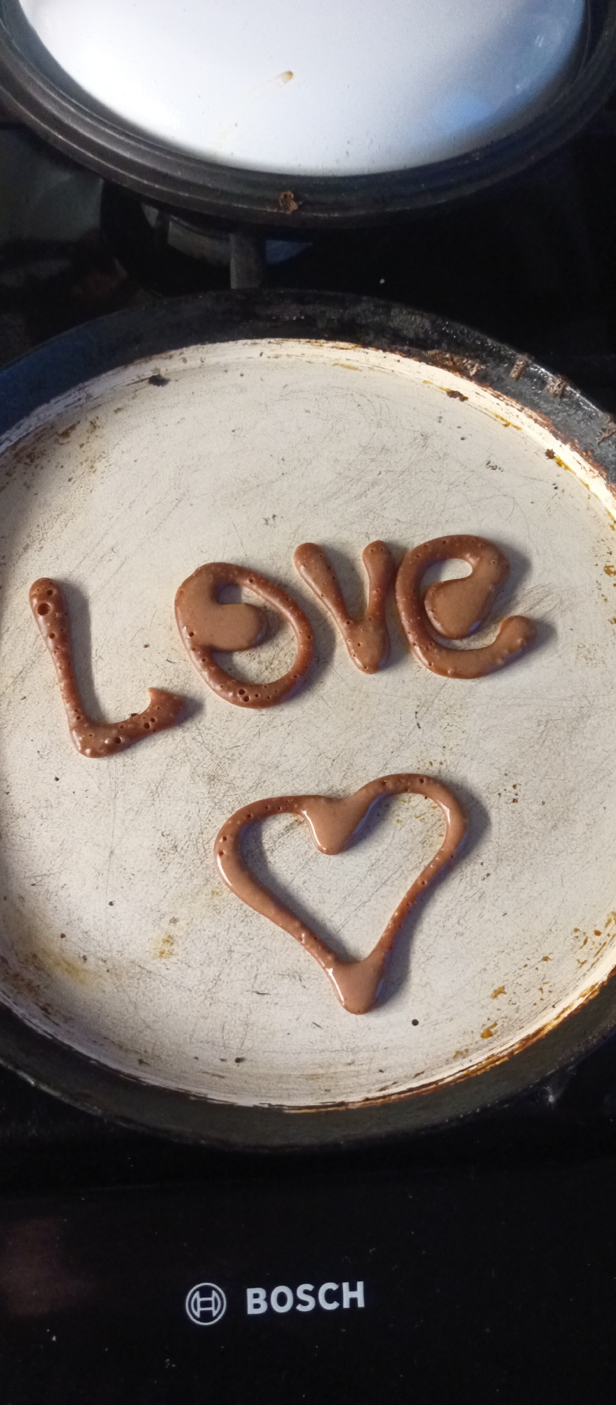 A high-angle shot depicts a white and slightly stained frying pan, containing the word "LOVE" spelled out in a brown, pancake-like batter, alongside a heart shape also formed from the same batter. The pan rests on a black Bosch-branded stovetop. The scene takes place indoors, potentially in a kitchen. No people are visible.