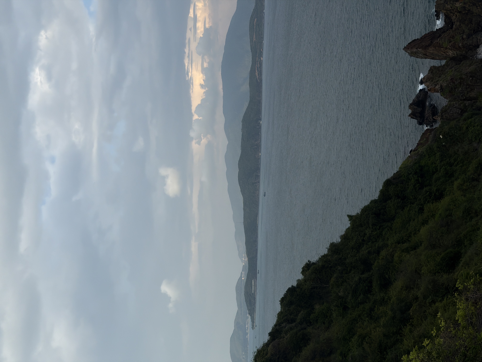 View of the coast from Nam Nha Trang Ward, Vietnam. The coastline is defined by layered mountains and a dark sea. Lush, green foliage covers the near foreground, while rocky outcroppings jut into the sea. The sky is overcast with diffuse clouds and hints of sunlight.
