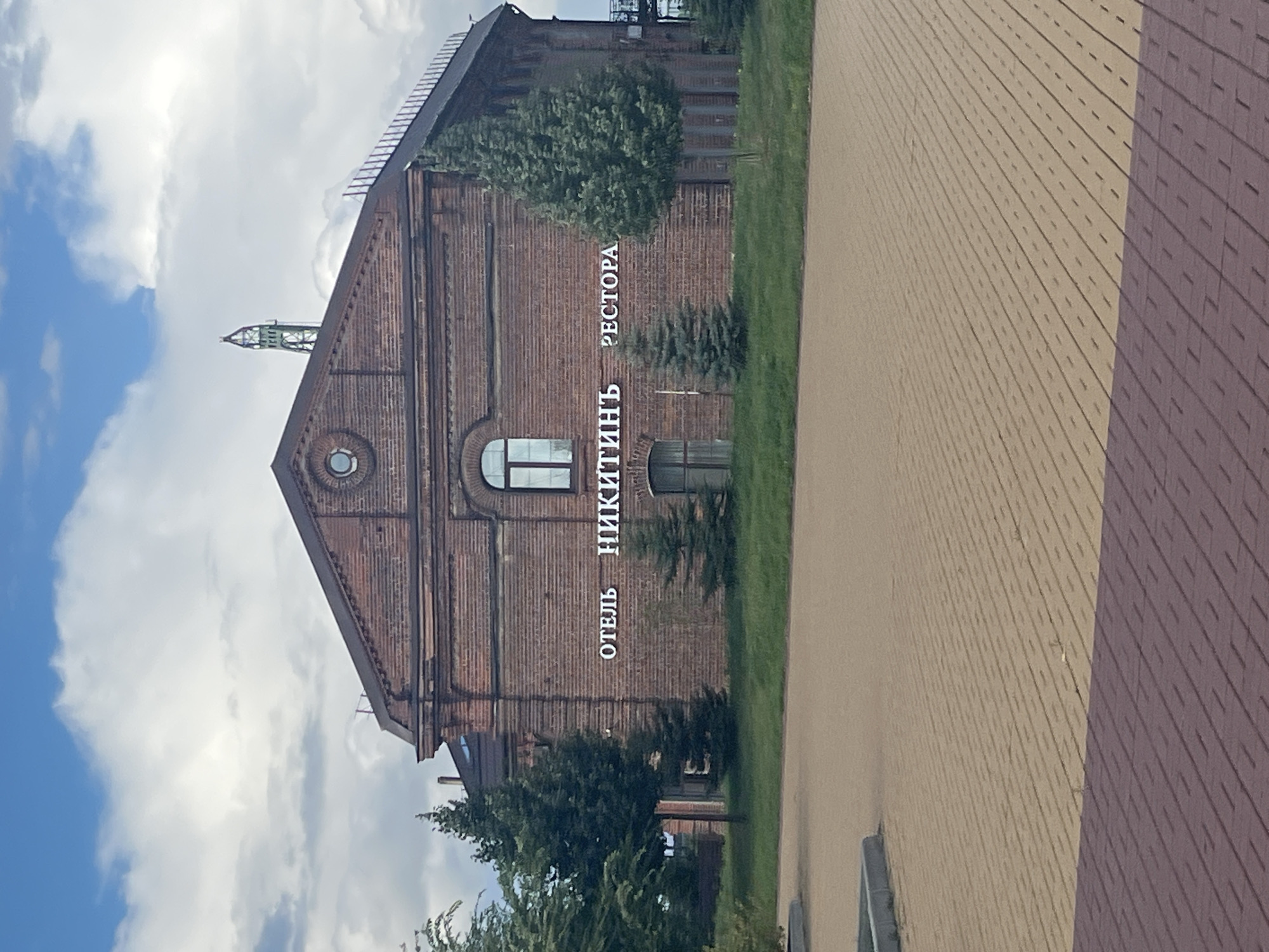 The image shows the exterior of a brick building with a sign that reads "ОТЕЛЬ НИКИТИНЪ РЕСТОРА". The building has arched windows and a triangular roof. There are trees in front of the building and a paved area in the foreground. The sky is cloudy. This building is located in Nizhny Novgorod, Russia. Specifically, it is Hotel Nikitin, a historic hotel in the city.