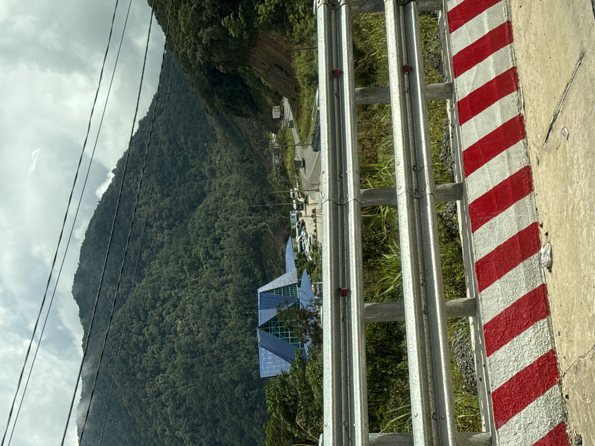 The image captures a scenic view of a mountainous landscape in Nam Khánh Vĩnh, Vietnam. In the foreground, a section of a road barrier with alternating red and white stripes is visible on the right side of the frame. The barrier appears to be made of metal. Beyond the barrier, lush green trees cover the steep slopes of a mountain. In the mid-ground, a winding road can be seen with several vehicles, including trucks and cars, traveling along it. Further into the valley, a distinct, modern building with a geometric, multi-faceted blue roof stands out against the natural backdrop. The sky is cloudy, suggesting an overcast day, and the overall atmosphere is serene and natural. The image is taken from a slightly elevated perspective, possibly from a car or a viewpoint overlooking the road and the valley. The orientation of the image is rotated 90 degrees counterclockwise from standard landscape orientation.