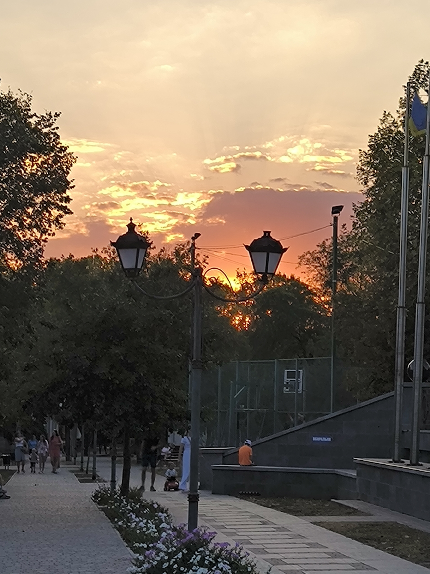 This image depicts a public park scene in Mykolaiv, Ukraine, during sunset. The sky is filled with dramatic clouds illuminated by the setting sun, casting warm hues of orange and yellow, with visible sun rays. In the foreground, a paved walkway lined with flowering bushes leads into the park. Two ornate street lamps are visible, one in the mid-ground and another further back. Trees line the walkway, creating a shaded canopy.
In the middle ground, there's a fence surrounding what appears to be a sports court, possibly for basketball, with a backboard visible. Several people are present in the park, including a group walking on the main path in the left mid-ground, and a child sitting on a toy car further down the path. A person in an orange shirt is seated on a low wall in the right mid-ground, facing a sports court area. Two flagpoles are visible on the right side of the image, one of which displays the Ukrainian flag. The overall impression is of a peaceful evening in a well-maintained urban park.