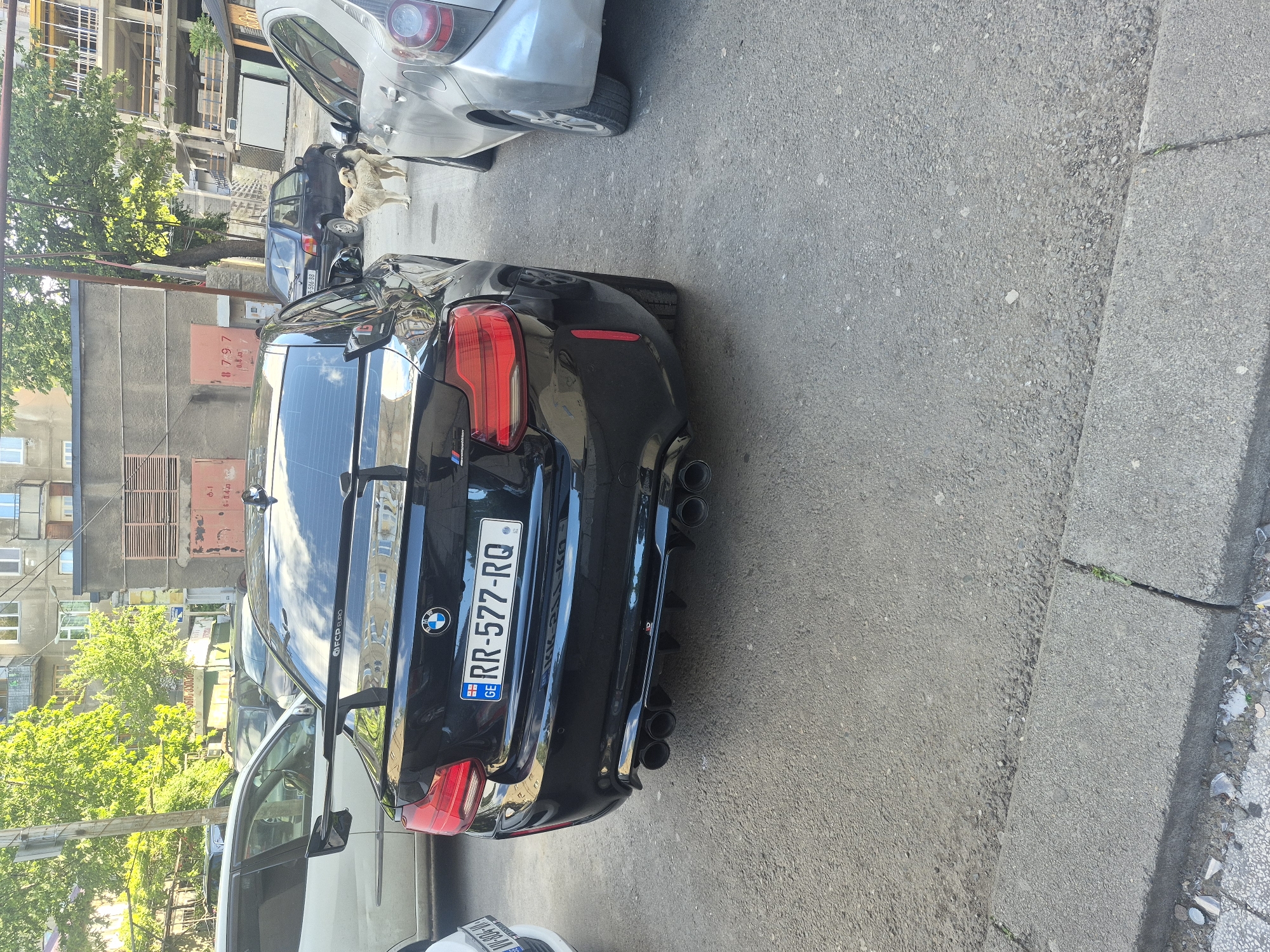 A black BMW is parked on a Tbilisi street with the license plate RR-577-RQ. The car is parked next to a curb and other parked vehicles. The background includes buildings, trees, and other urban elements.