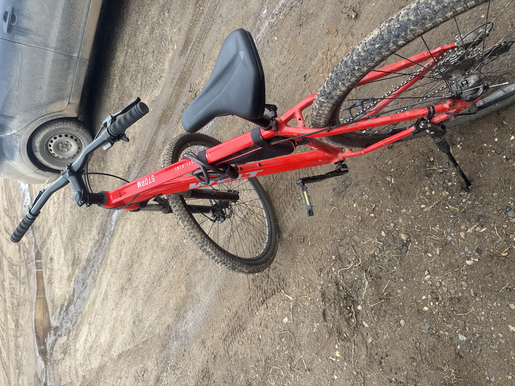 The image shows a red mountain bike lying on muddy ground next to a car. The bike is a "STORM" model. The location appears to be outdoors, possibly an unpaved area or dirt road. The background shows vegetation and general outdoor detritus scattered on the muddy ground. There are no people visible in the frame. The location is Кемуль, Russia.