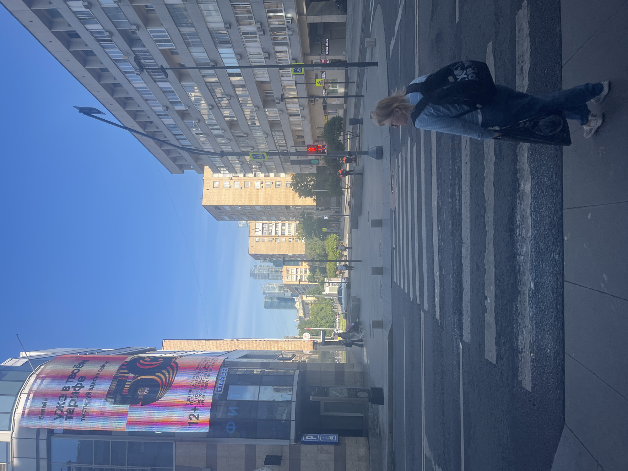 The image depicts a bright, sunny city street scene from an elevated perspective. A wide road with a prominent zebra crossing extends through the center, flanked by sidewalks and buildings.
On the right side of the frame, a woman with blonde hair stands on the sidewalk next to the pedestrian crossing. She is wearing a light blue long-sleeved top, blue jeans, and sneakers, with a black backpack slung over her shoulders. She holds a dark, flat personal mobility device, possibly a foldable electric scooter or board, resting on the ground. A traffic light displaying a red pedestrian signal is visible on the opposite side of the crossing, suggesting she is waiting to cross.
To the left, a tall, multi-story building with numerous windows and balconies lines the street. In the background, a clear blue sky spans above a cityscape featuring various buildings and some greenery.
In the mid-ground on the right, a modern building features a large, colorful LED billboard. The billboard displays text in Cyrillic script, including "Билайн" (Beeline), "УЖЕ В ТВОЁМ ТАРИФЕ" (Already in your tariff), "тестируй здесь и сейчас" (test here and now), and a "12+" age restriction. Other legible texts on nearby signs include "MAER," "OZON," and "АТЕЛЬЕ" (Atelier). Various street signs, including parking signs and warning signs, are also visible. The clear sky and bright lighting indicate it is daytime. The Cyrillic script confirms the location context of Moscow, Russia.