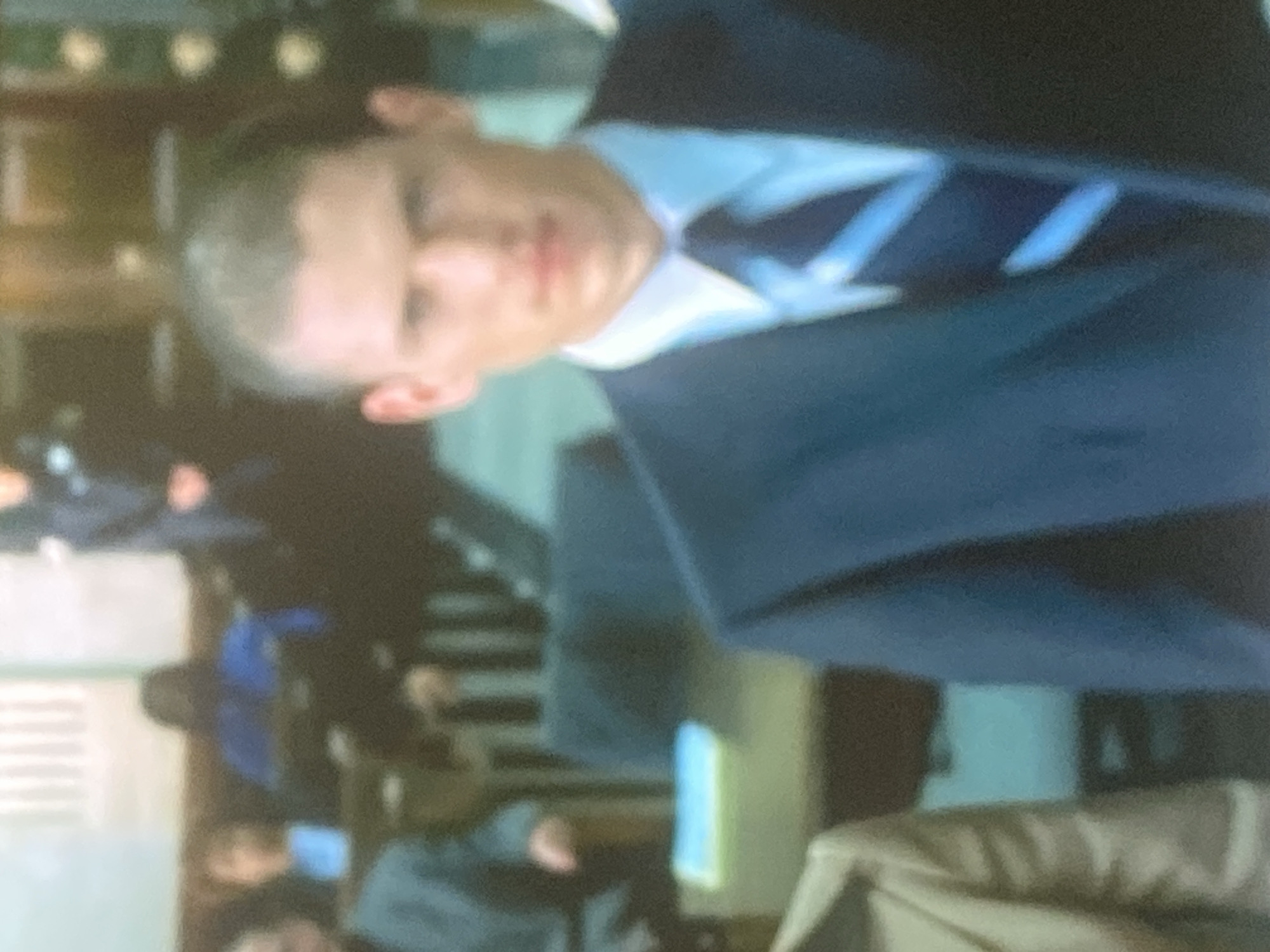 The image features a young man with short, light-colored hair, appearing to look directly at the viewer. He is formally dressed in a dark suit jacket, a light blue collared shirt, and a blue and white diagonal striped tie. His head is slightly tilted.
The scene is an indoor setting, and the background is heavily blurred, making specific details indistinct. However, it appears to contain several other blurred figures, some of whom are wearing blue clothing, potentially uniforms. Hints of furniture, possibly chairs or tables, and structural elements like walls or windows are vaguely visible. The overall ambiance suggests a formal gathering, such as a meeting, a class, or an institutional event. The man's expression is neutral.
The image quality is low, characterized by blurriness and pixelation, particularly in the background. There is no visible text within the frame. While the provided context suggests the location is Gosa, Nigeria, there are no specific visual cues in the image itself to corroborate this geographical detail.