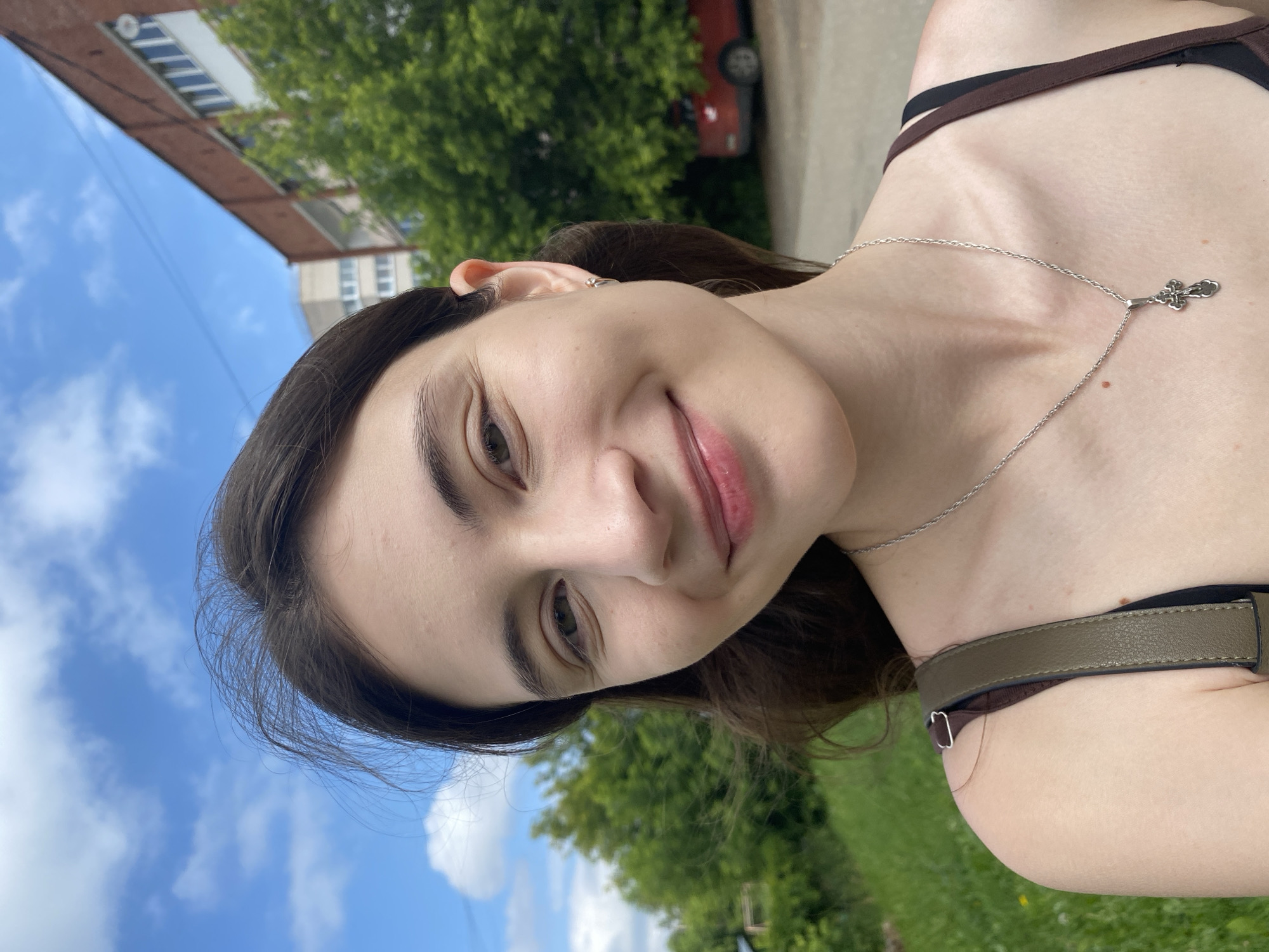 A young woman with long, dark brown hair is taking a selfie outdoors. She has a light complexion, brown eyes, and is smiling. She is wearing a silver necklace with a cross pendant and a dark-colored top with visible straps. Her left ear has a small silver earring. The background suggests an urban or suburban environment in Votkinsk, Russia, with a brick building and green trees visible. The sky is blue with scattered clouds, and there is a red car parked in the background. The image is taken from the chest up, with the woman's face being the primary focus.