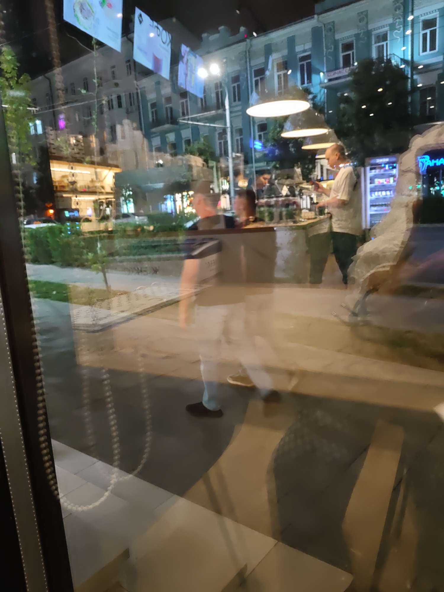 The image captures a reflection on a glass surface, likely a storefront window, in Rostov-on-Don, Russia. The scene depicts people walking past a building at night, illuminated by artificial lighting. In the foreground, a man in a white t-shirt and dark pants is visible, holding a phone and facing away from the camera. He appears to be standing near a counter with bottles and glasses. Further back, another person in a dark shirt and light pants is walking. The background shows a building with multiple windows and balconies, and some greenery. The reflection also reveals posters or advertisements hanging above. The overall impression is a street scene at night with activity in what appears to be a cafe or restaurant.