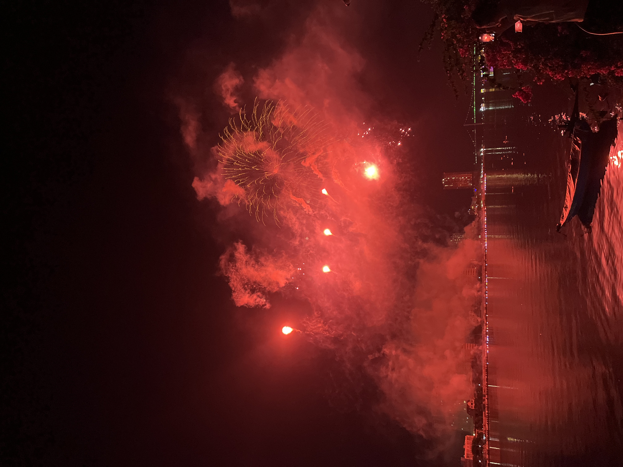 The image captures a nighttime fireworks display over the Han River in the city of Đà Nẵng, Vietnam. The scene is dominated by vibrant red and orange light from the fireworks and associated smoke, which casts a crimson glow across the sky and reflects intensely on the water's surface.
In the upper two-thirds of the frame, multiple fireworks explode, creating large plumes of red smoke and bursts of golden-yellow light. Several bright, concentrated points of light, indicating active firework launches or smaller explosions, are visible amidst the smoke.
The lower third of the image shows the calm surface of the Han River, reflecting the intense red light of the fireworks and the illuminated city buildings. Ripples on the water distort some of the reflections. In the immediate foreground on the right, a person is seen from the chest up, holding a smartphone with both hands, positioned to record or photograph the fireworks. Red-hued foliage, possibly flowering bushes, partially obscures the lower right corner, extending into the water. A small, dark-hulled boat with a lighter-colored interior or stripe is moored near the riverbank in the lower-left foreground, partially illuminated by the red light.
Across the river, the city skyline is visible. A prominent, tall skyscraper, identifiable as the Novotel Danang Premier Han River, stands on the opposite bank in the mid-ground. To its right, a cable-stayed or suspension bridge spans the river, adorned with lights that are also reflected in the water. Numerous smaller buildings and lights delineate the city's riverfront along the horizon.
