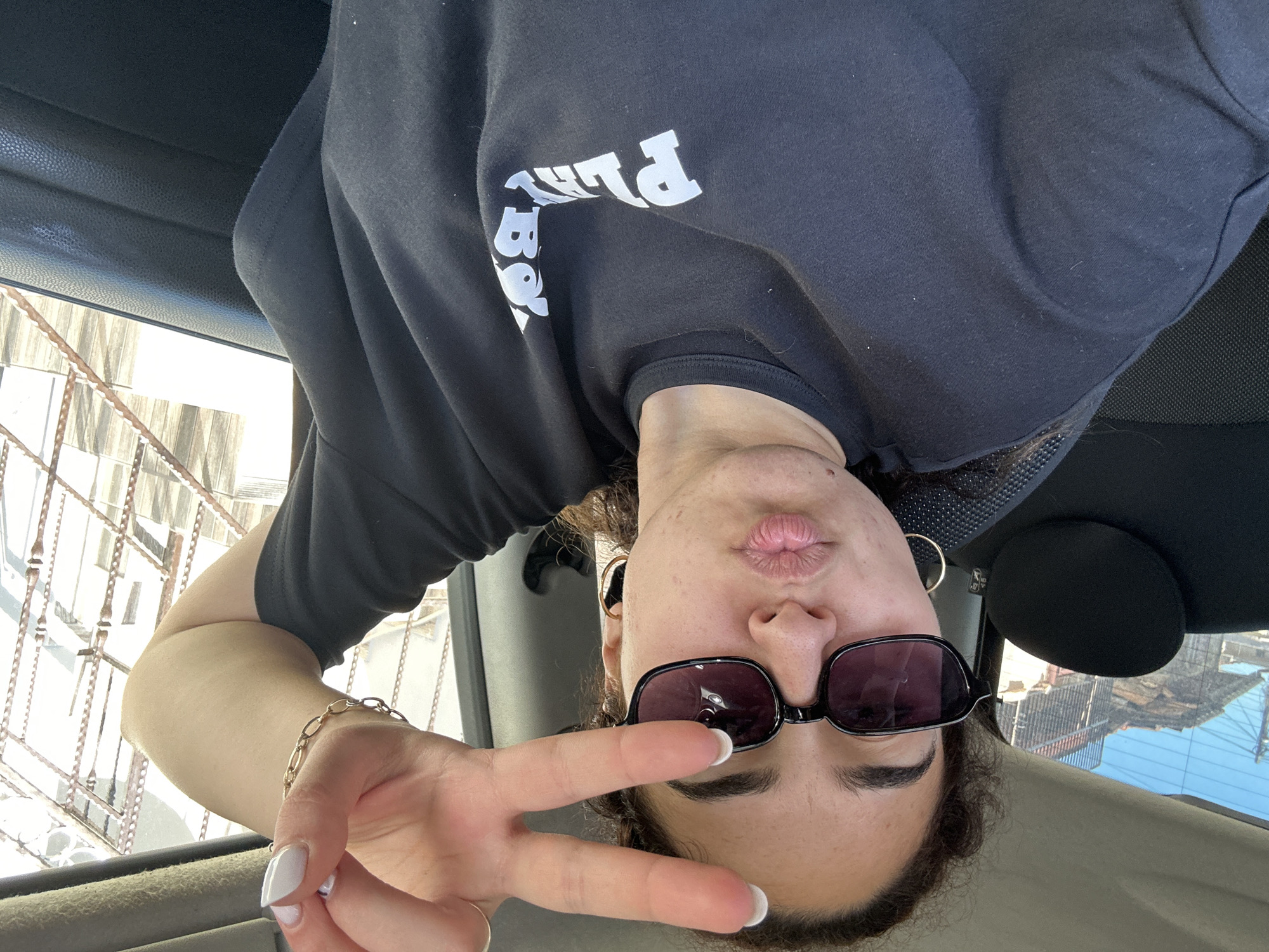 A woman is in a car, oriented upside down in the frame, wearing a black t-shirt with white lettering that says "Play Boy". She is wearing sunglasses and gold hoop earrings, holding up a hand showing a peace sign. She has a gold bracelet. The interior of the car is visible, including the ceiling and possibly a seat. Outside the car window, there appears to be a building under construction or renovation, possibly with scaffolding. Based on the style of architecture and the limited visual context, the image does not provide enough information to pinpoint the precise location in Burgas, Bulgaria.