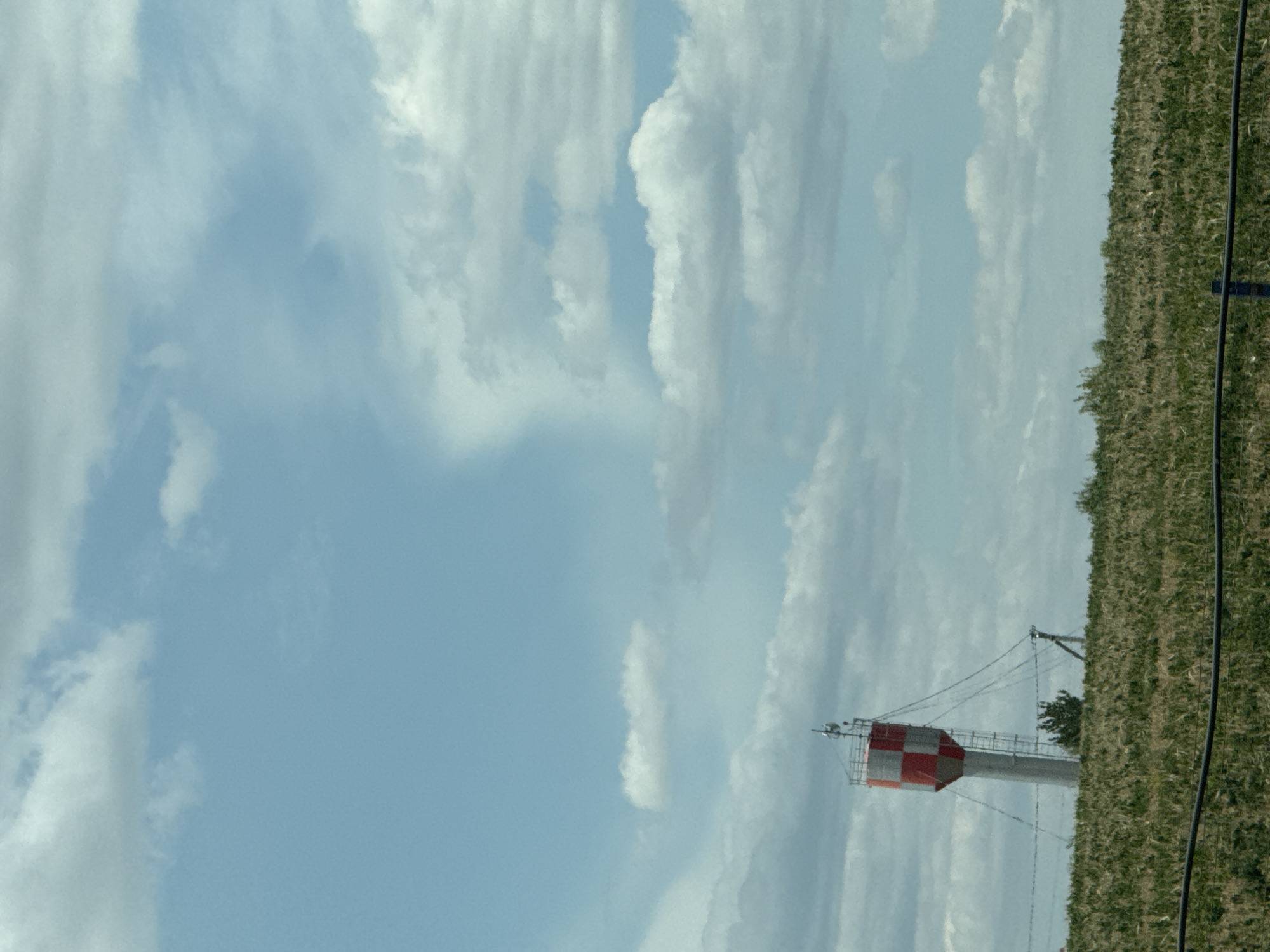 The image shows a water tower located in Horesti, Moldova. The water tower is cylindrical with a conical top, painted with a red and white checkered pattern. It is situated on a grassy hill, with a line of trees in the background. A ladder is attached to the side of the tower, and wires extend from the top of the tower to poles on the ground. The sky above is blue with scattered white clouds. The foreground of the image shows a field of green grass and a dark horizontal line, possibly a fence or cable.