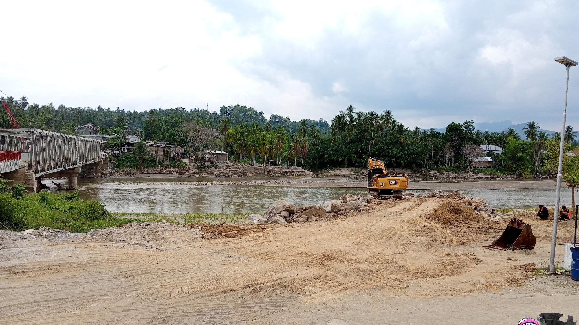 The image depicts a scene near Beunyot, Indonesia, characterized by a construction or repair project along a river. A large, grey metal bridge is visible on the left, spanning the muddy, brown river. The riverbanks are lined with lush green vegetation and palm trees, with a cluster of buildings situated behind the trees.
In the foreground, a construction site is evident. A yellow excavator, branded with "CAT", is positioned on a dirt path. Piles of dirt and large rocks are scattered around, suggesting ongoing earthmoving activities. A detached excavator bucket lies on the ground near the construction area. Two individuals, wearing dark clothing, are seated near a lamppost on the right side of the image, observing the area. The sky is overcast with grey clouds. The overall scene conveys a sense of infrastructure work in a tropical, rural setting.