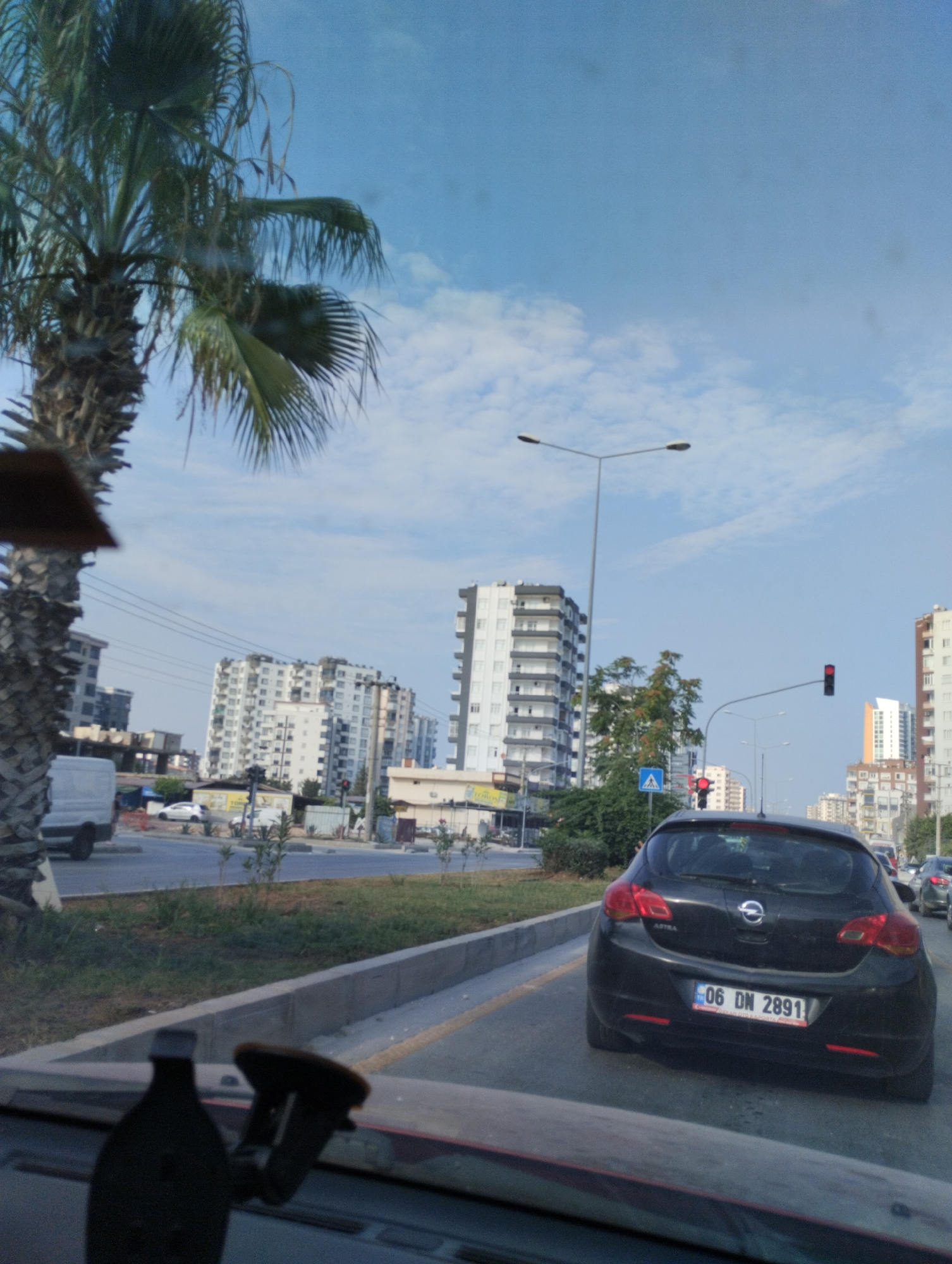 A black Opel Astra with license plate 06 DN 2891 is parked on a street in Mezitli, Turkey. The car is in the foreground, facing away from the camera. In the background, there are several tall buildings, a palm tree, and a street lamp. The sky is blue with a few white clouds. The overall scene appears to be a typical urban environment.