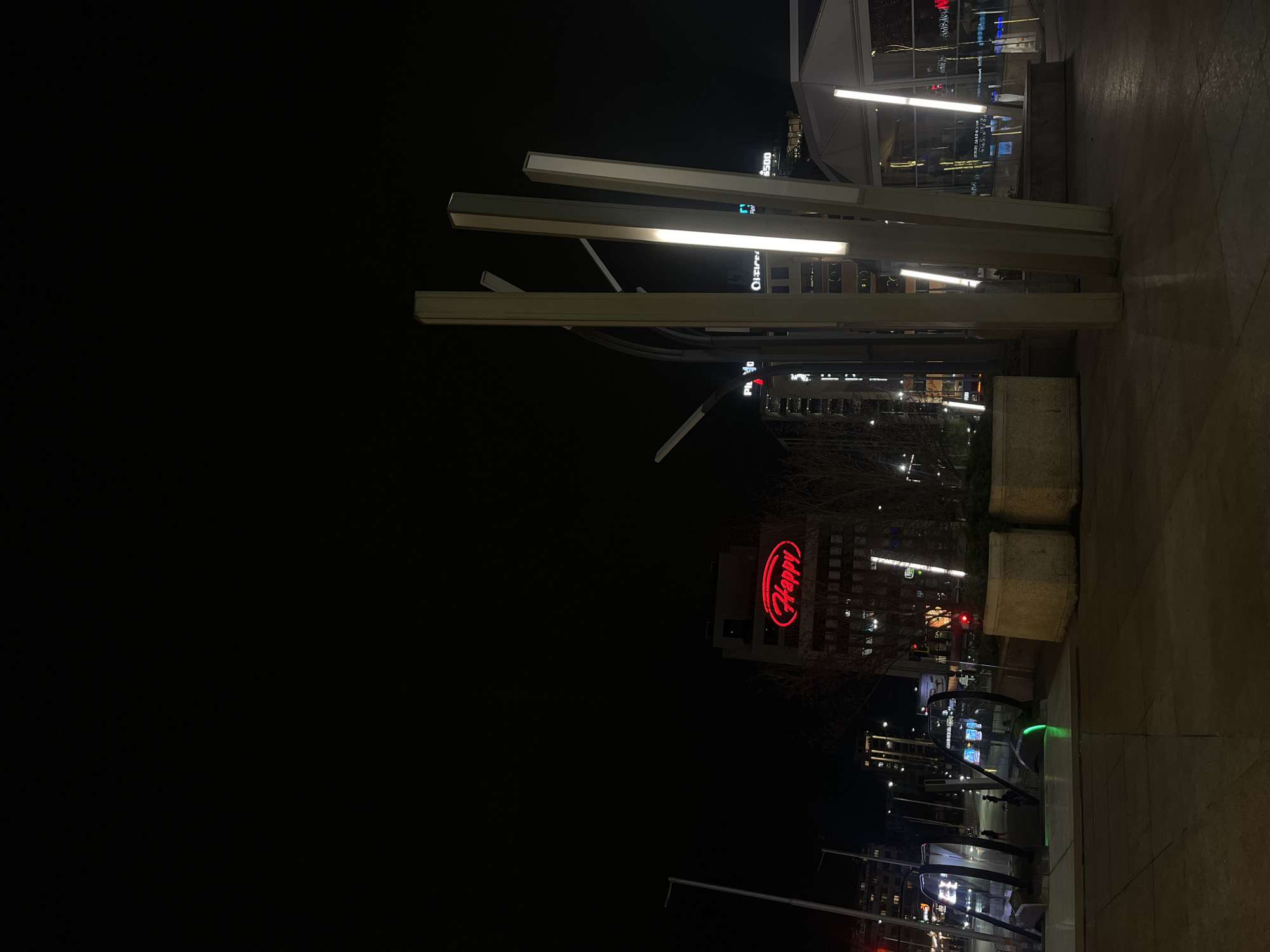 The image depicts a night scene in Sofia, Bulgaria, with a prominent "Happy" restaurant sign illuminated in red on a building in the background. In the foreground, a modern architectural structure with illuminated white bars stands on a paved surface. Pedestrian barriers are visible on the right, and a building with multiple illuminated windows is visible beyond the structure. The scene is dark, with artificial lighting highlighting key elements.