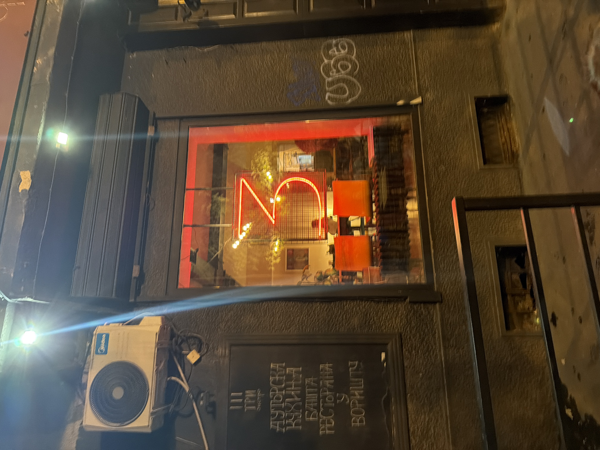 The image depicts the exterior of a building at night, likely a restaurant or bar, located in Belgrade, Serbia. A large window with a red neon sign shaped like the number "3" is prominently featured. Inside the establishment, tables and chairs are visible, illuminated by warm lighting. To the left of the window, a dark chalkboard displays text in Cyrillic script. Above the window, an air conditioning unit is mounted on the wall. The surrounding building facade is rough-textured and dark. Graffiti is visible on the wall to the right of the window. The ground in front of the building appears wet, suggesting recent rain. A metal railing runs along the bottom of the image, and a small vent is visible in the wall below the window.