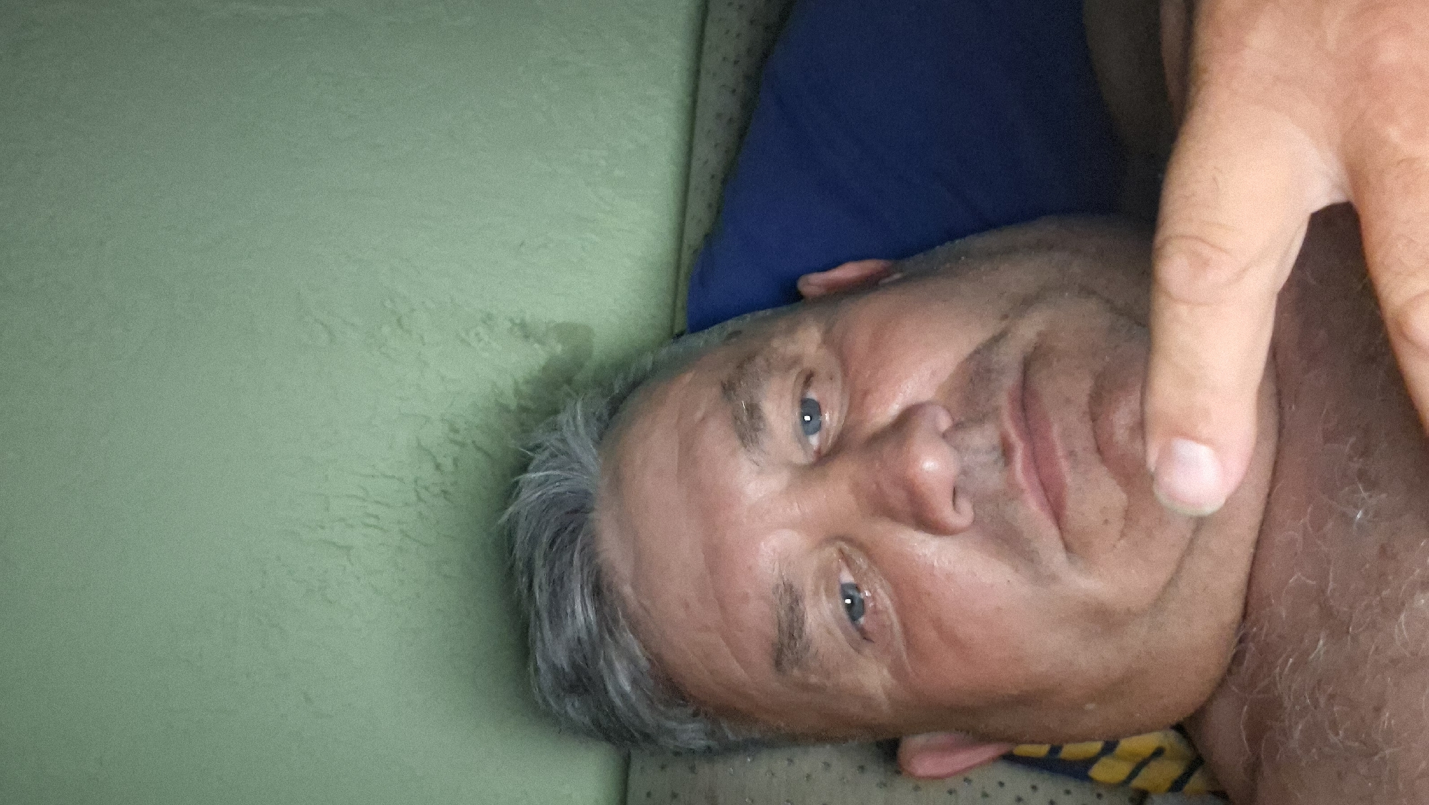 The image is a close-up, selfie-style photograph of an adult male. He has graying hair, light blue eyes, and appears to be lying down. His head is tilted slightly, and his chin is resting on his hand. A blue pillow is visible behind him. The background wall is a light green color. The image is lit from the front, likely by a camera flash. There is no discernible indication of the city of Voinova, Moldova, or any specific urban or rural setting within the image. The focus is solely on the individual's face and upper body.