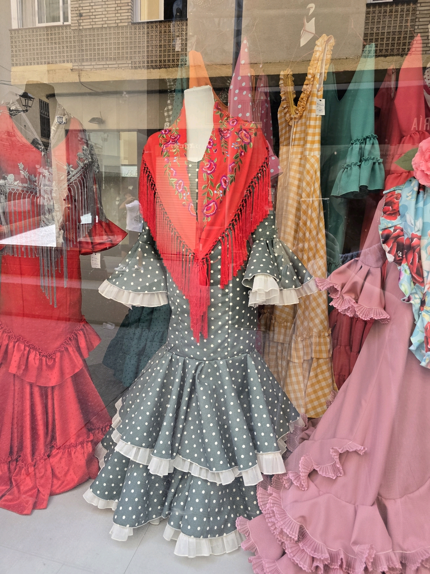 This is a medium-shot photograph taken through a glass storefront, showcasing a display of flamenco dresses. The dresses are vibrantly colored and feature traditional flamenco styling, with ruffled skirts and decorative bodices. One dress in the center is a dark grey with white polka dots, adorned with a red shawl featuring floral embroidery and red fringe. To its left, a bright red flamenco dress with a ruffled hem is visible. To the right, a pale pink flamenco dress with multiple layers of ruffled trim is displayed. In the background, other dresses in various colors and patterns, including yellow gingham and teal, can be seen. Reflections in the glass suggest the dresses are on mannequins and the scene is likely a shop. The backdrop appears to be an urban setting with building facades visible through the reflection, hinting at a street-level display in a city. Given the style of the dresses, this establishment is likely a flamenco costume shop in or around Jaén, Spain, known for its flamenco heritage.