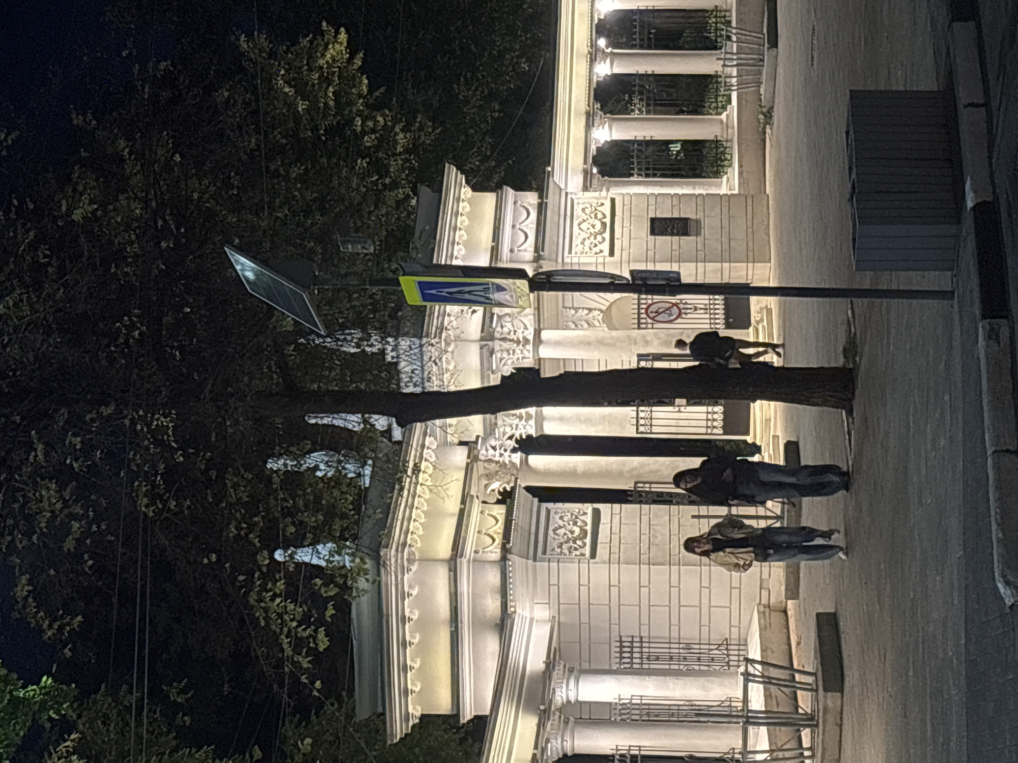 The image depicts an outdoor scene at night, likely in the city of Novaya Usman', Russia. In the foreground, a paved walkway is visible, with a decorative concrete planter to the right. The walkway leads towards an ornate white stone building with columns, decorative moldings, and wrought-iron fencing. Three individuals are walking on the walkway. Two women are walking together towards the left of the frame, and a man is walking towards the right, away from the camera. A traffic sign with a pedestrian crossing symbol is mounted on a pole in the middle ground, near the building entrance. Trees with dark foliage are present in the background, obscuring the sky. A solar panel is mounted on a pole near the traffic sign. The overall lighting suggests artificial illumination from the building and streetlights.
