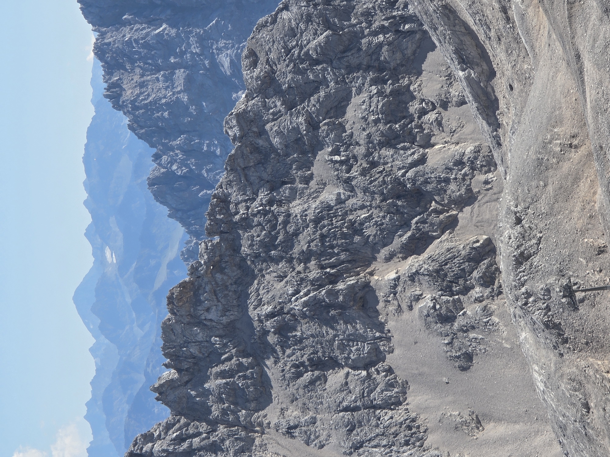 The image displays a view of the Zugspitze mountain range, Germany's highest peak, as seen from a high vantage point. The foreground features rugged, rocky terrain with visible scree slopes and rock formations. In the mid-ground, a ridge of gray, weathered rock stretches across the frame, characterized by sharp peaks and crevices. Beyond this ridge, a series of progressively distant mountain ranges are visible, fading into a hazy blue due to atmospheric perspective. The sky above is clear and light blue with a few wispy clouds. There are no people or specific structures identifiable in the image, focusing solely on the natural mountainous landscape. The environment is clearly alpine and mountainous.